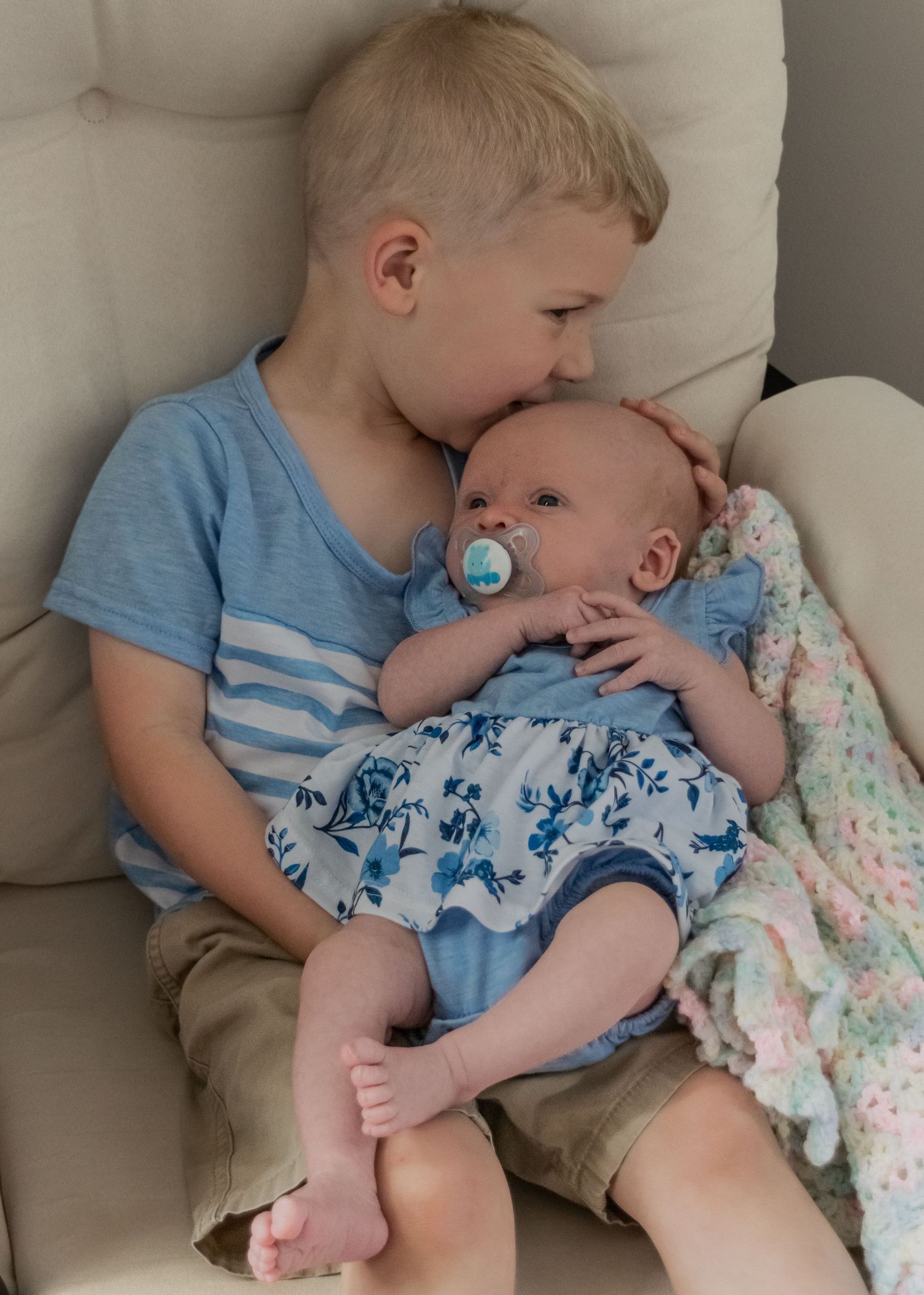 Young blonde boy kissing newborn sibling with pacifier while sitting together in gray chair.