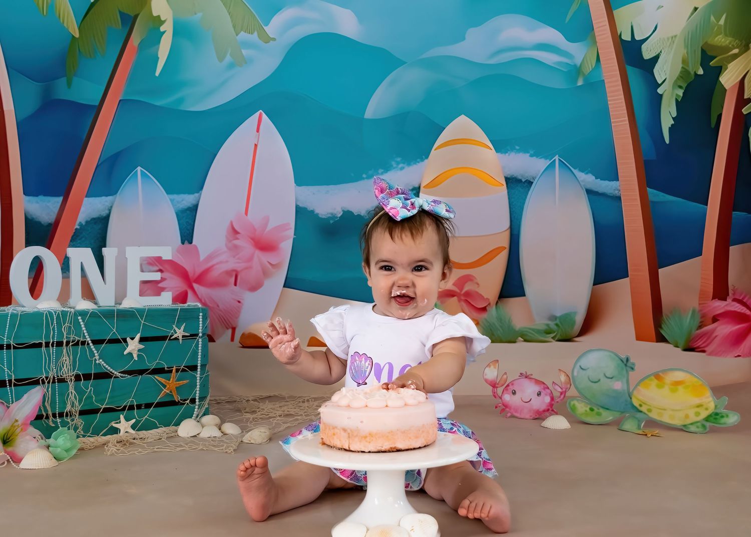 Smiling baby girl in white outfit reaches toward pink cake at tropical beach-themed first birthday smash.