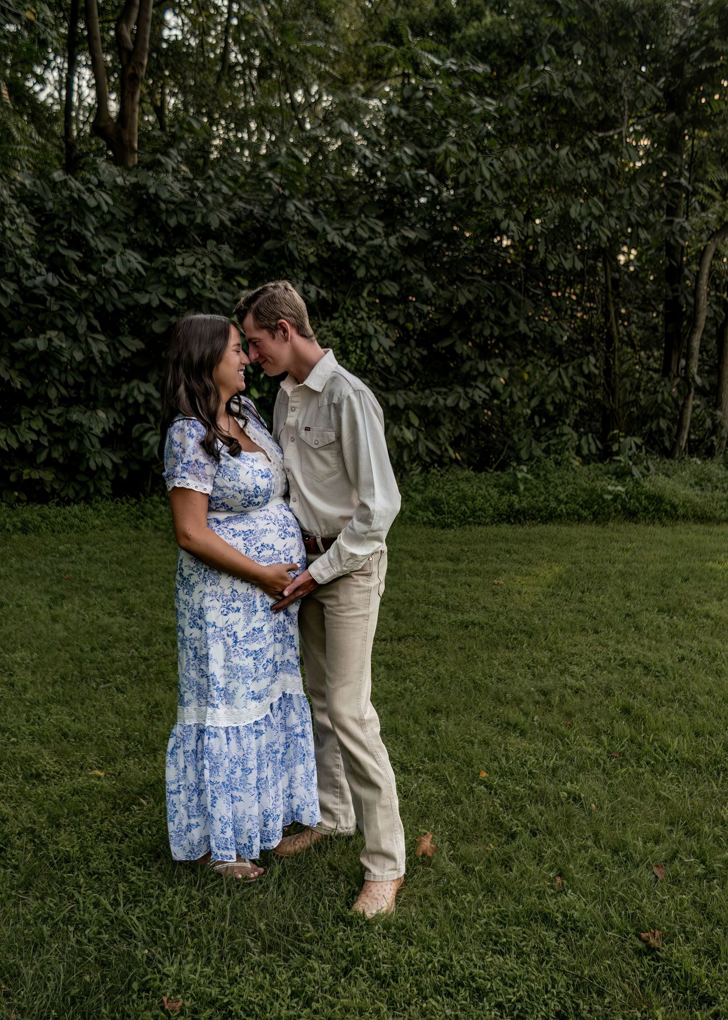 Pregnant couple in blue floral and beige outfits sharing tender moment on green lawn.