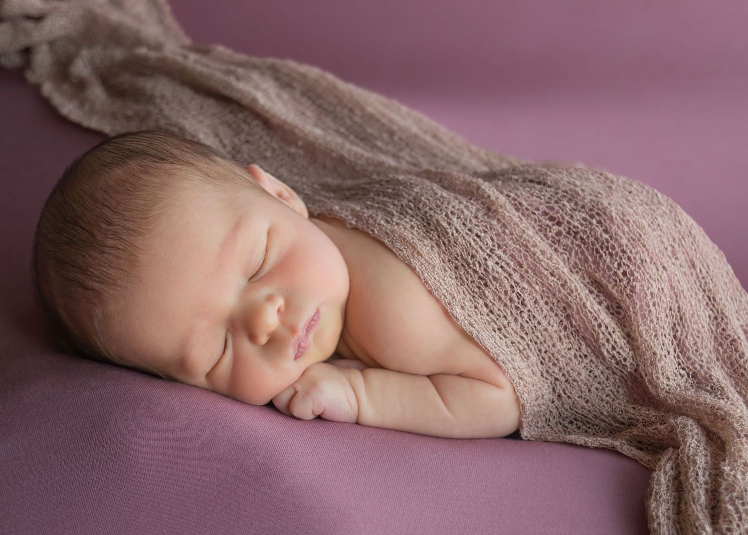 Sleeping newborn baby wrapped in a soft pink knit blanket on a purple background.