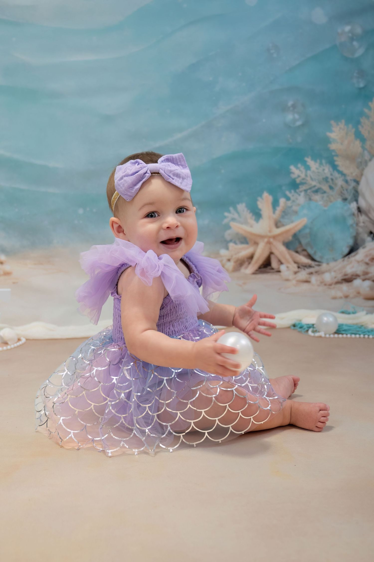 Baby girl in purple mermaid dress celebrating her first birthday with ocean backdrop.