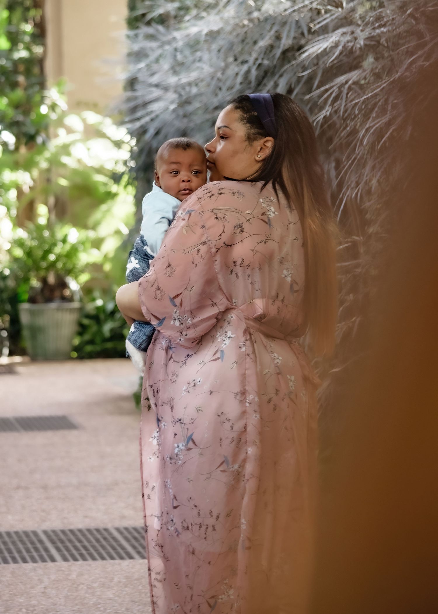 Mother in pink floral dress kissing her baby boy while standing near tropical plants outdoors.