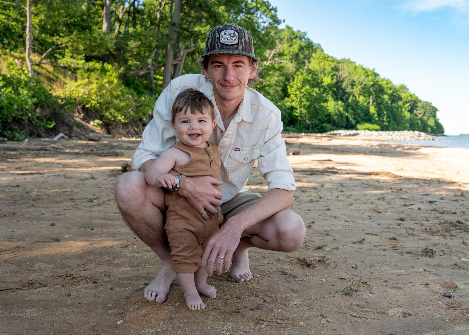 Young man crouching on sandy beach with toddler boy, lush green trees in background.