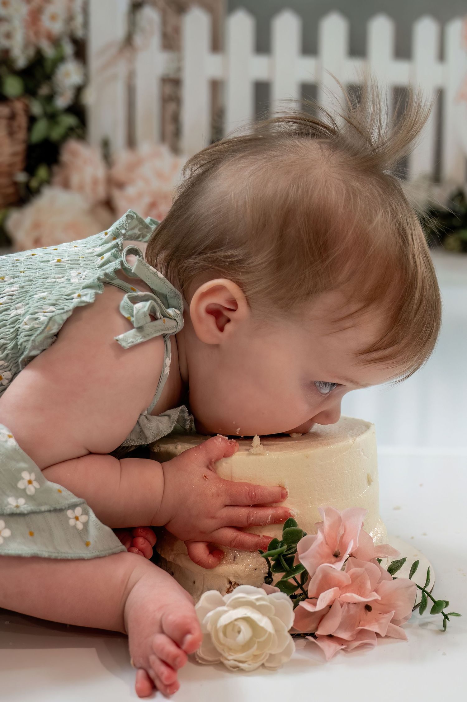 Baby girl in sage dress leans in to bite first birthday smash cake surrounded by pink roses.