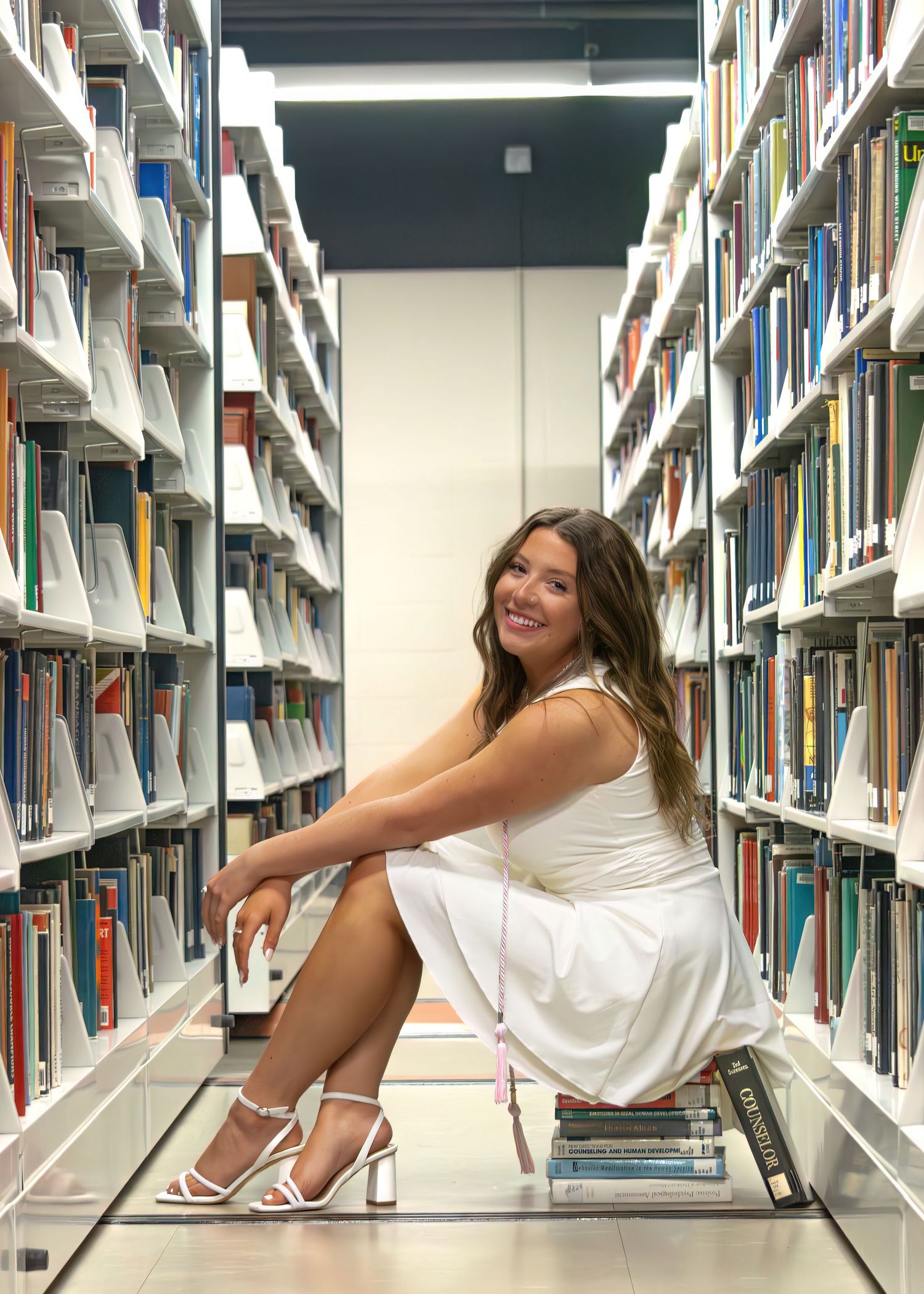 A brunette woman in a white dress sits smiling between tall bookshelves in a library aisle.