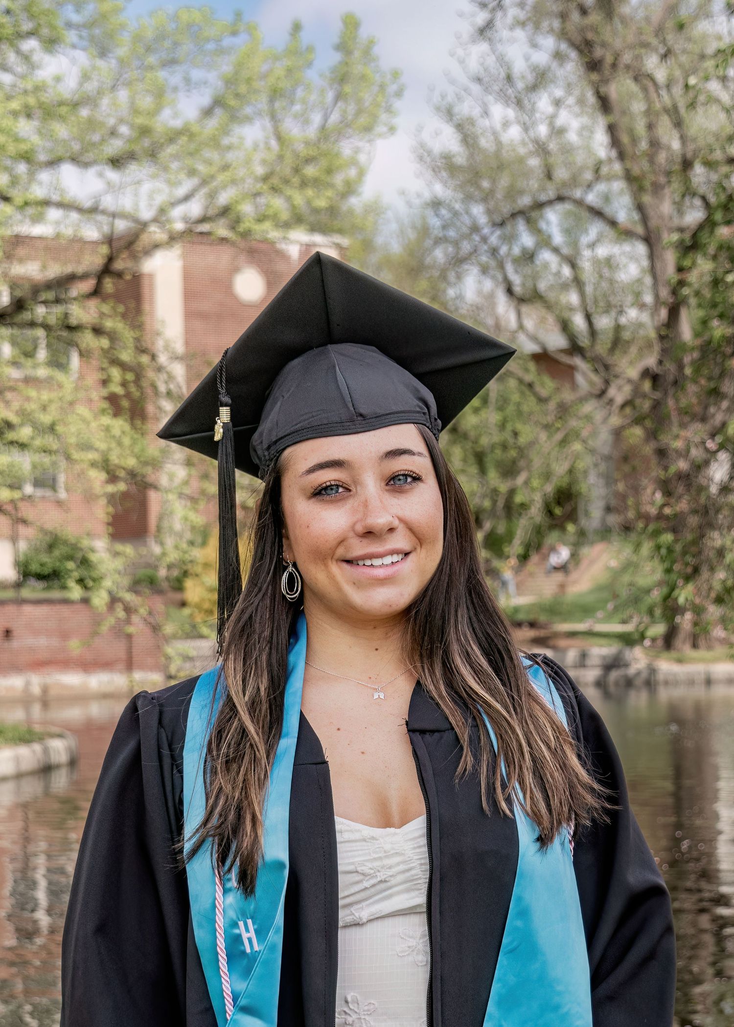 Smiling female graduate in black cap and teal graduation stole posing outdoors near campus pond.