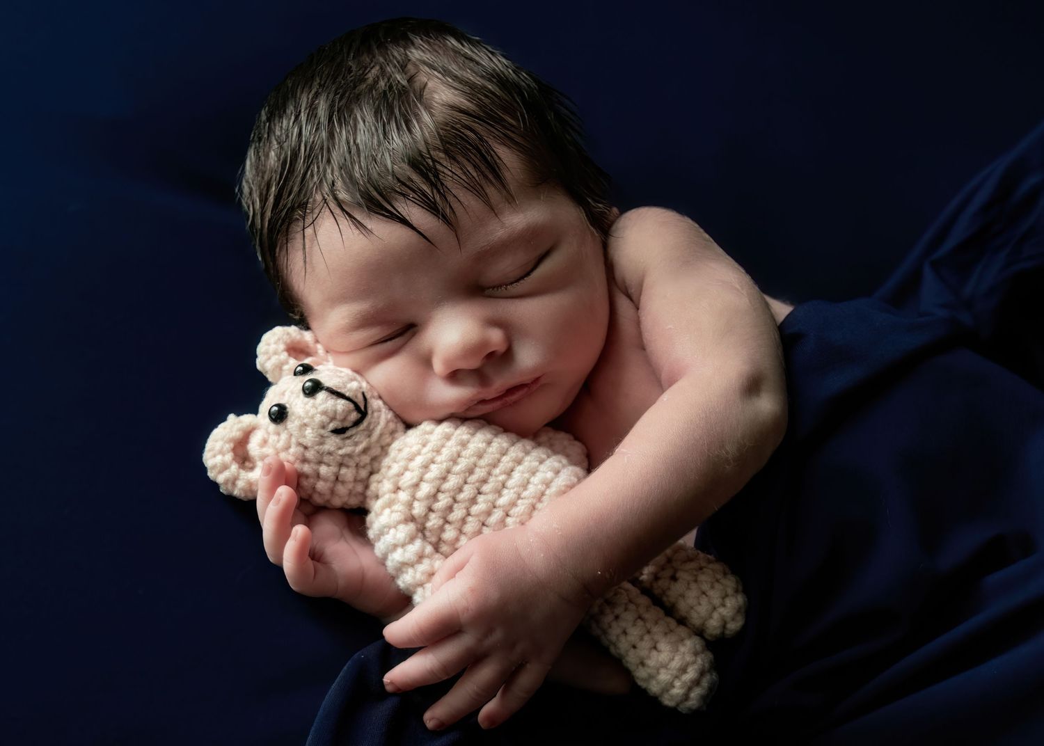 Peaceful newborn baby sleeping while hugging a cream knit teddy bear on navy blue backdrop.