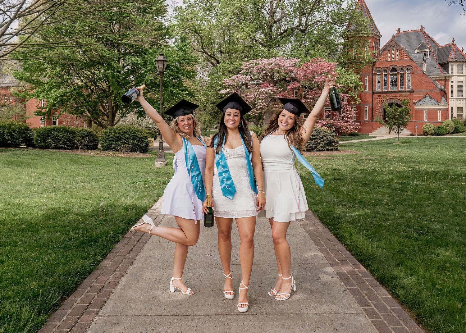 Three graduates in white dresses and caps celebrate on a campus walkway surrounded by spring blooms.
