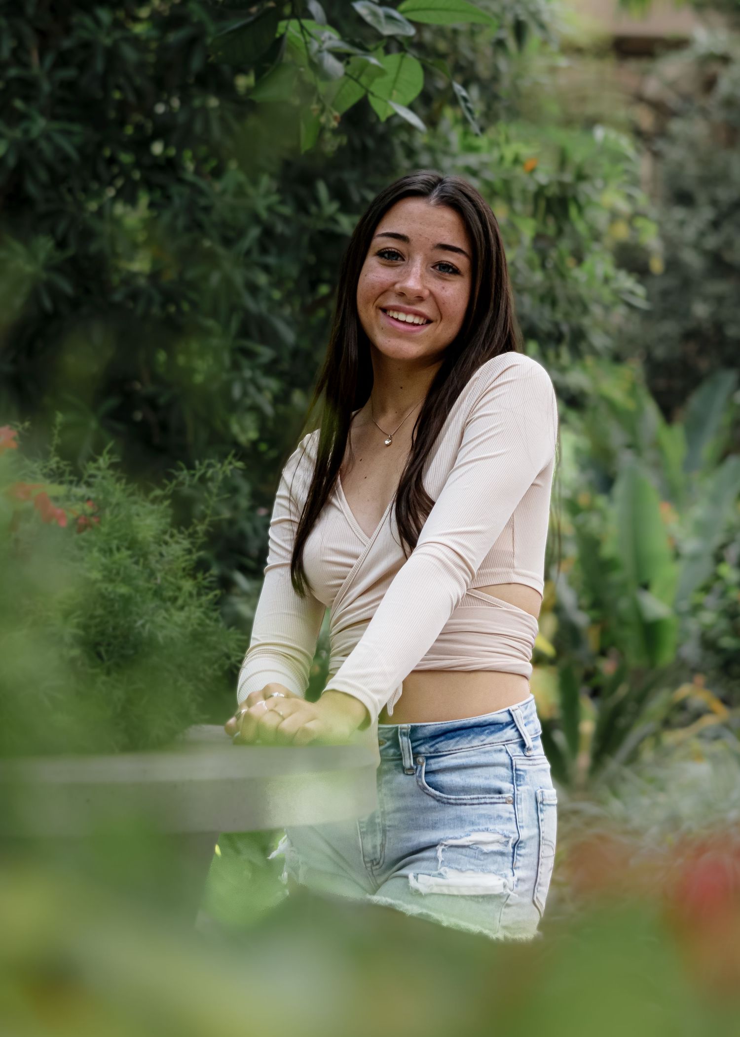 Young woman in beige crop top and denim shorts smiling while gardening among lush green plants.