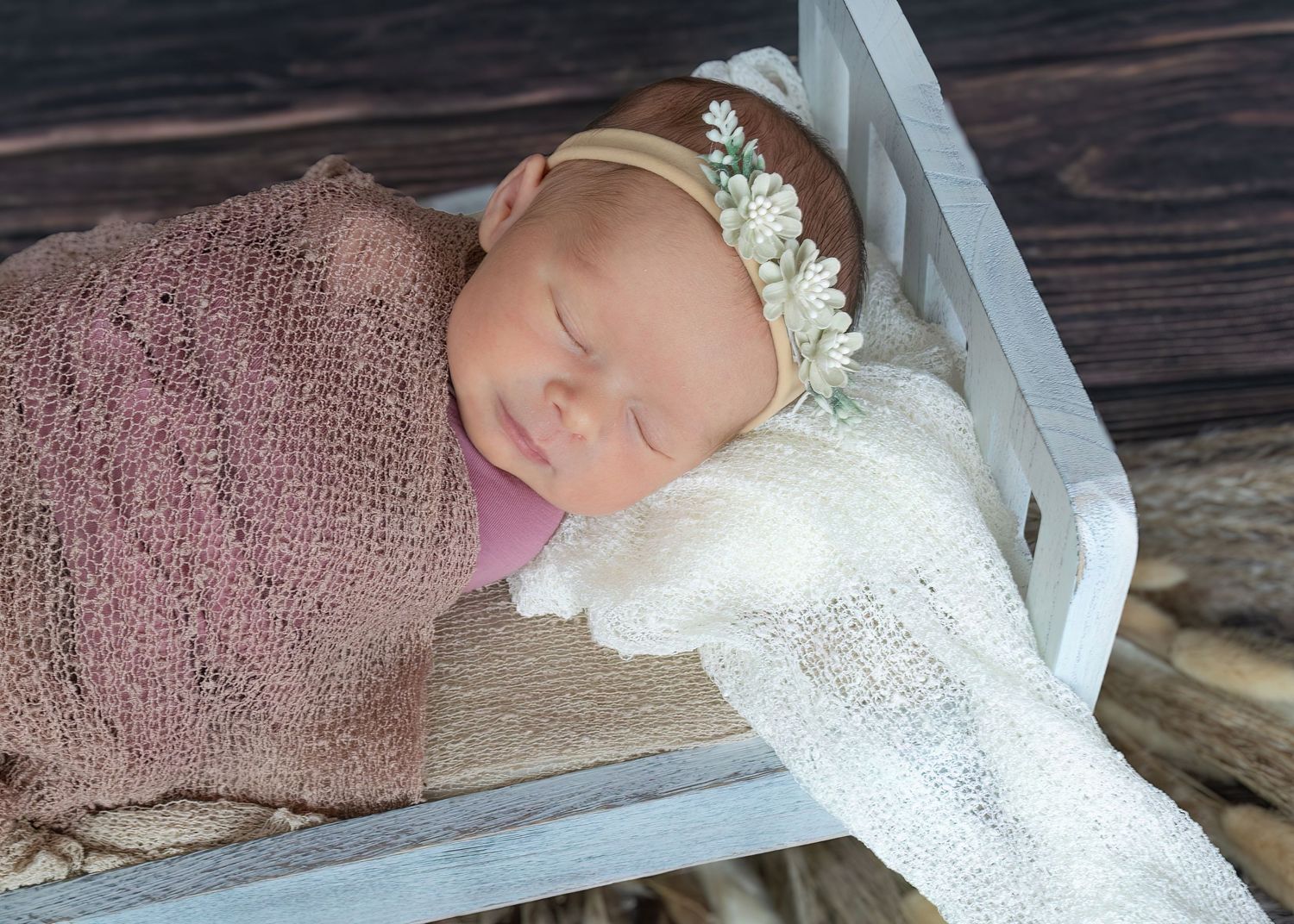 Sleeping newborn baby wrapped in pink knit swaddle with floral headband in white wooden bed.