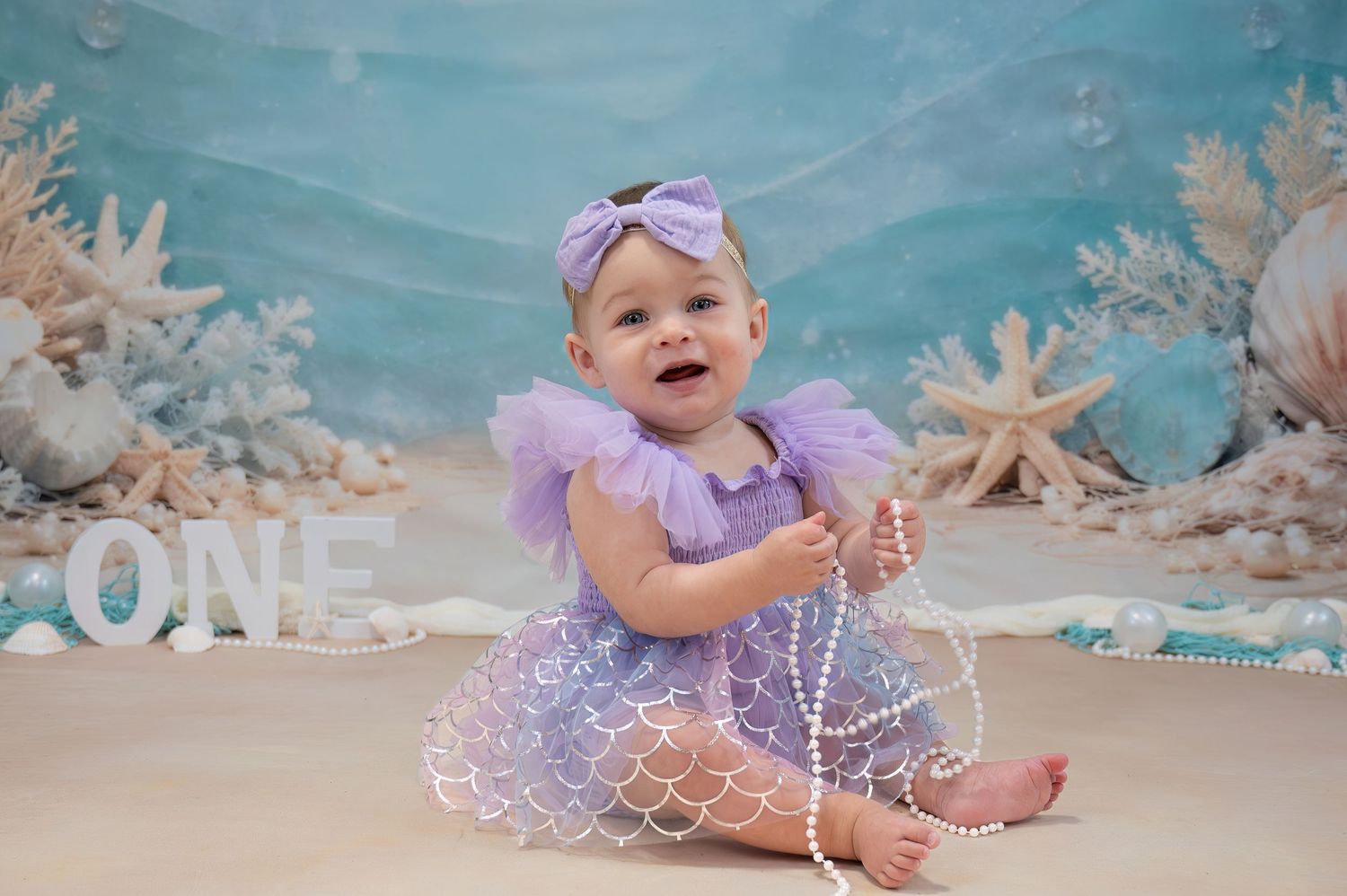 Baby girl in purple mermaid dress celebrates first birthday with ocean backdrop.