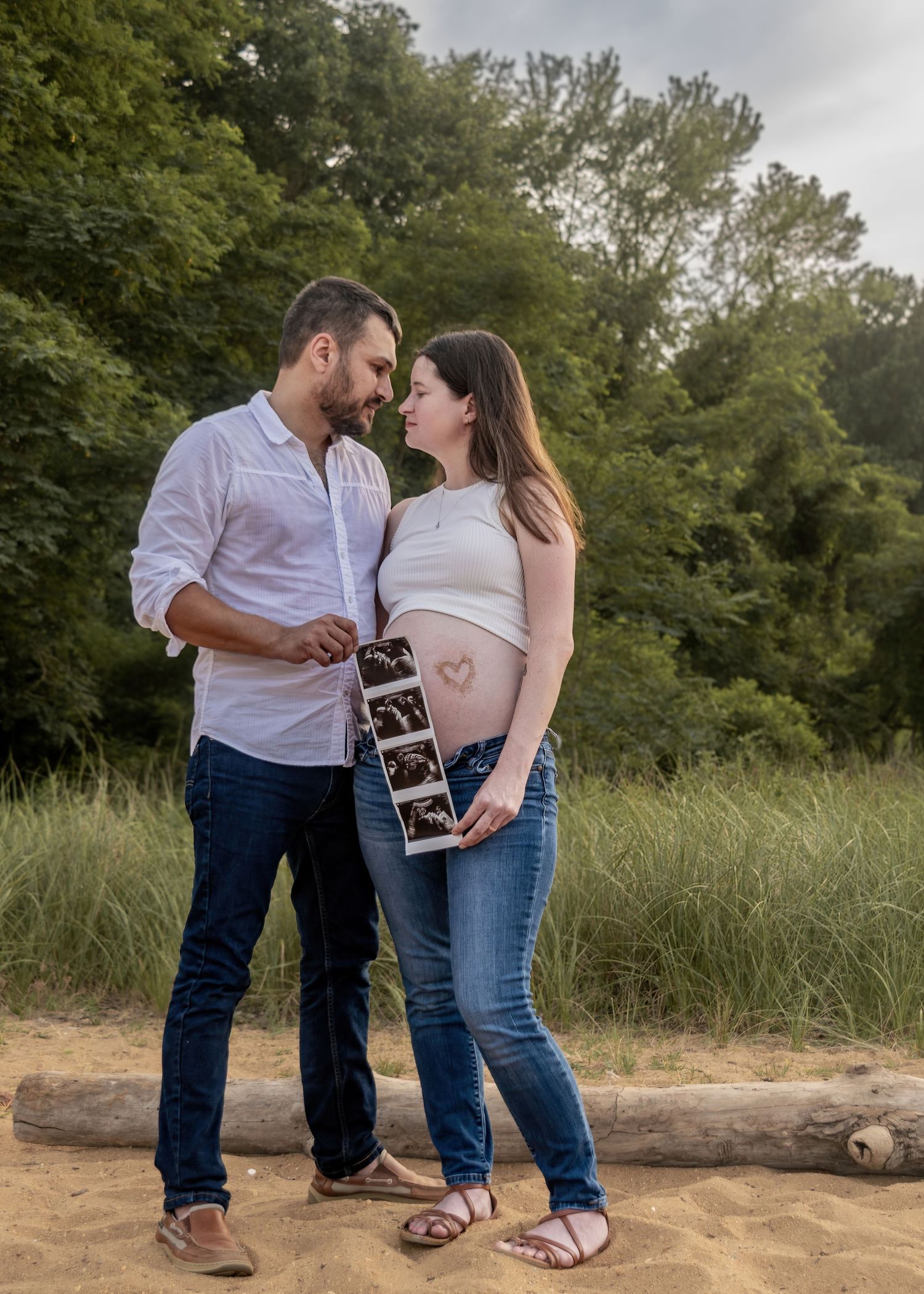 Pregnant couple holding ultrasound photos outdoors on sandy beach with lush green trees behind them.