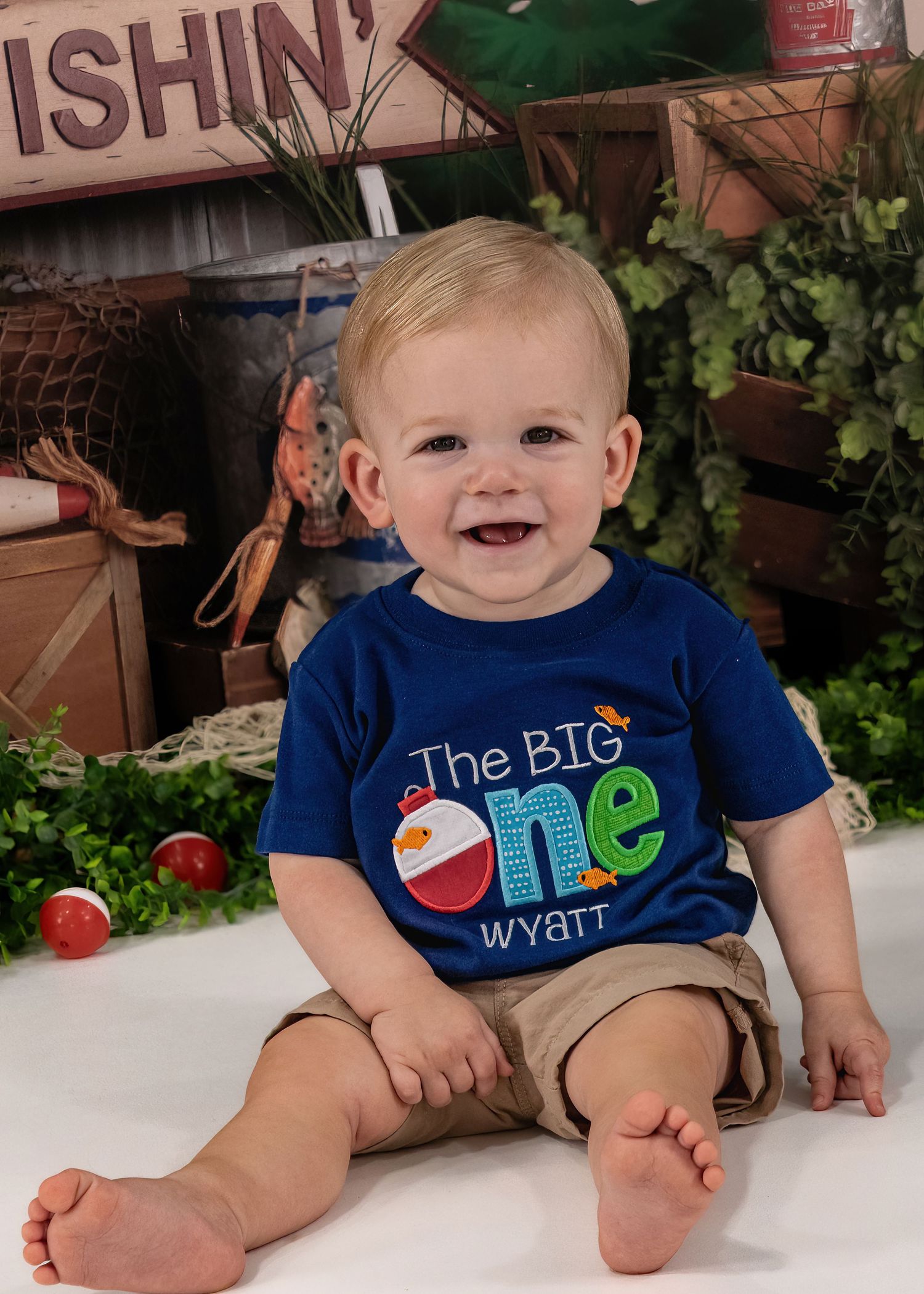 Baby boy Wyatt smiles in blue shirt reading The Big One for his fishing-themed first birthday photos.