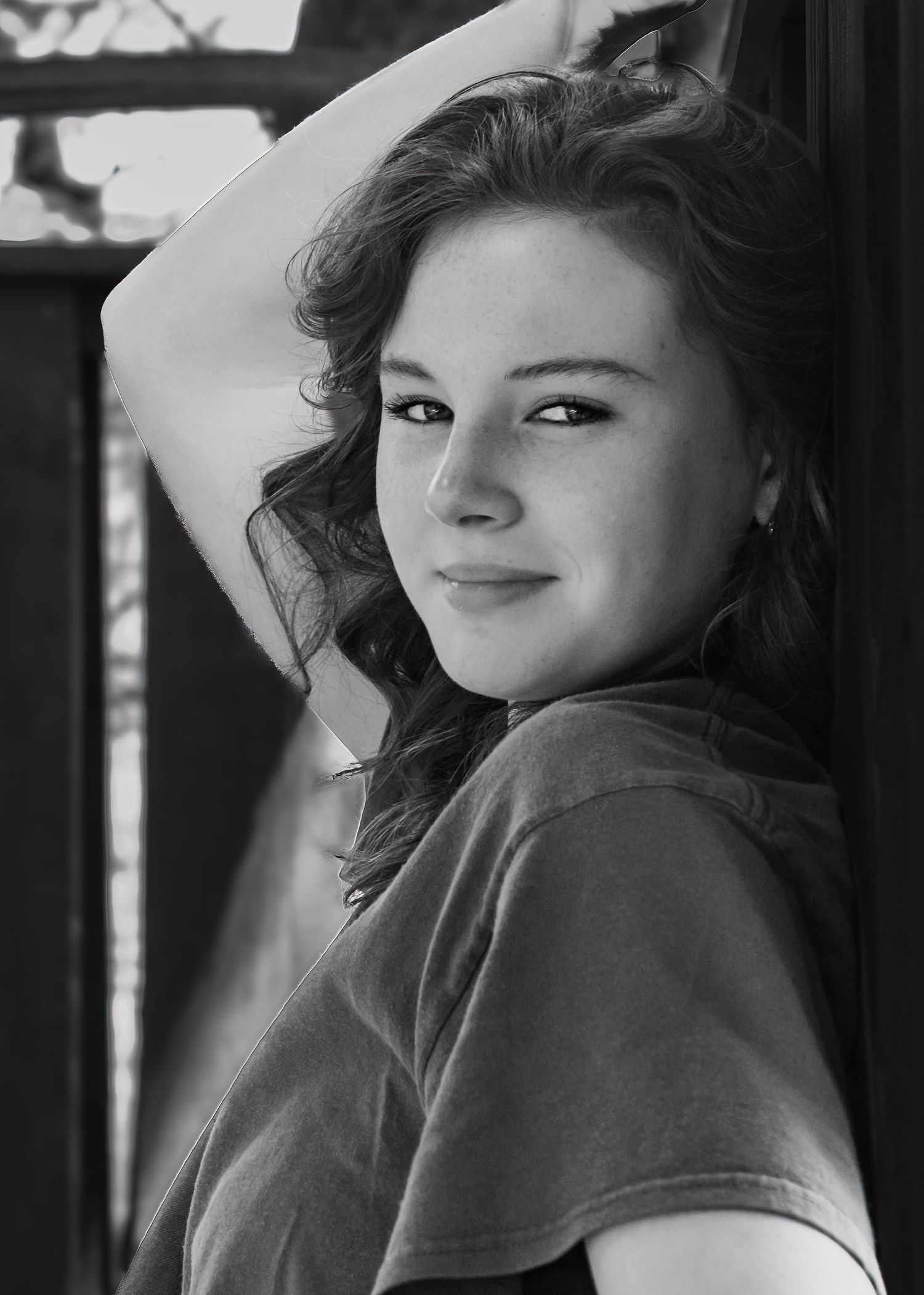 Black and white portrait of a young woman with wavy hair leaning against a wall with her arm raised.