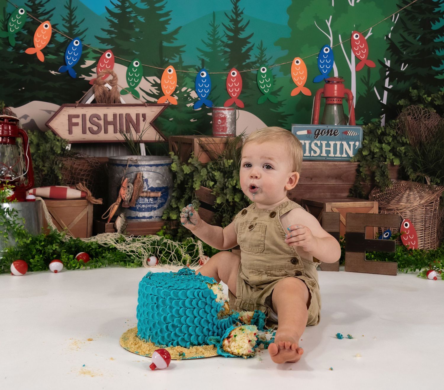 Curious baby in khaki overalls explores smashed blue cake at fishing-themed first birthday photo session.