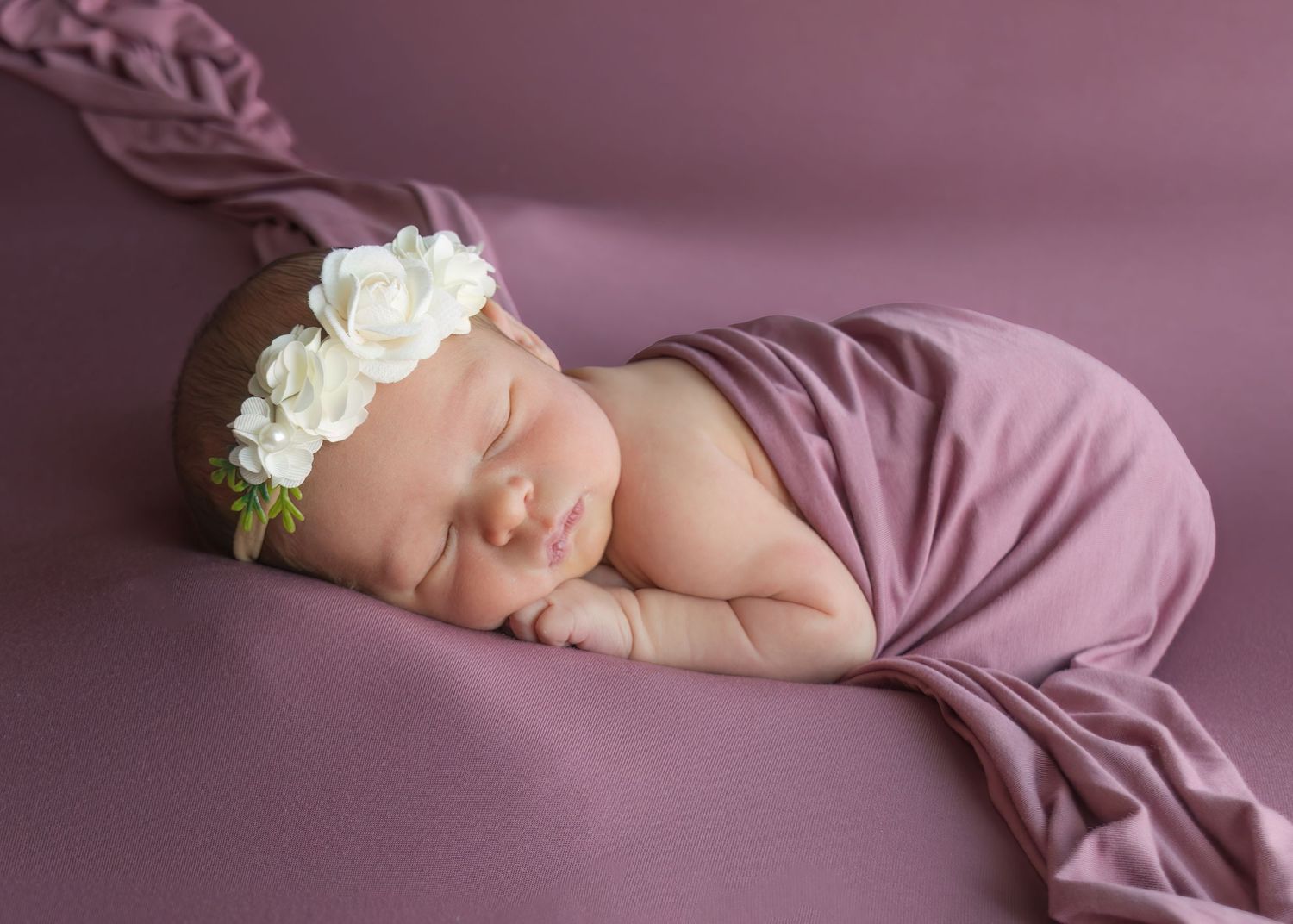 Peaceful newborn baby sleeping on mauve purple wrap with white rose floral headband.