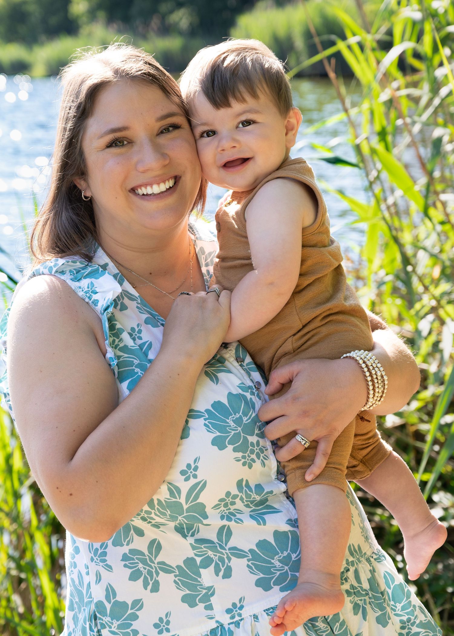 Smiling mother holds happy baby boy outdoors near a sunlit river.