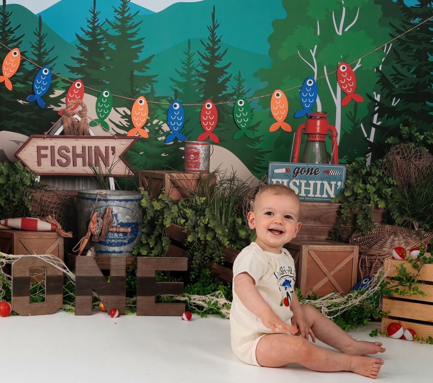 Happy baby boy sits smiling at colorful fishing-themed first birthday cake smash photography session outdoors.