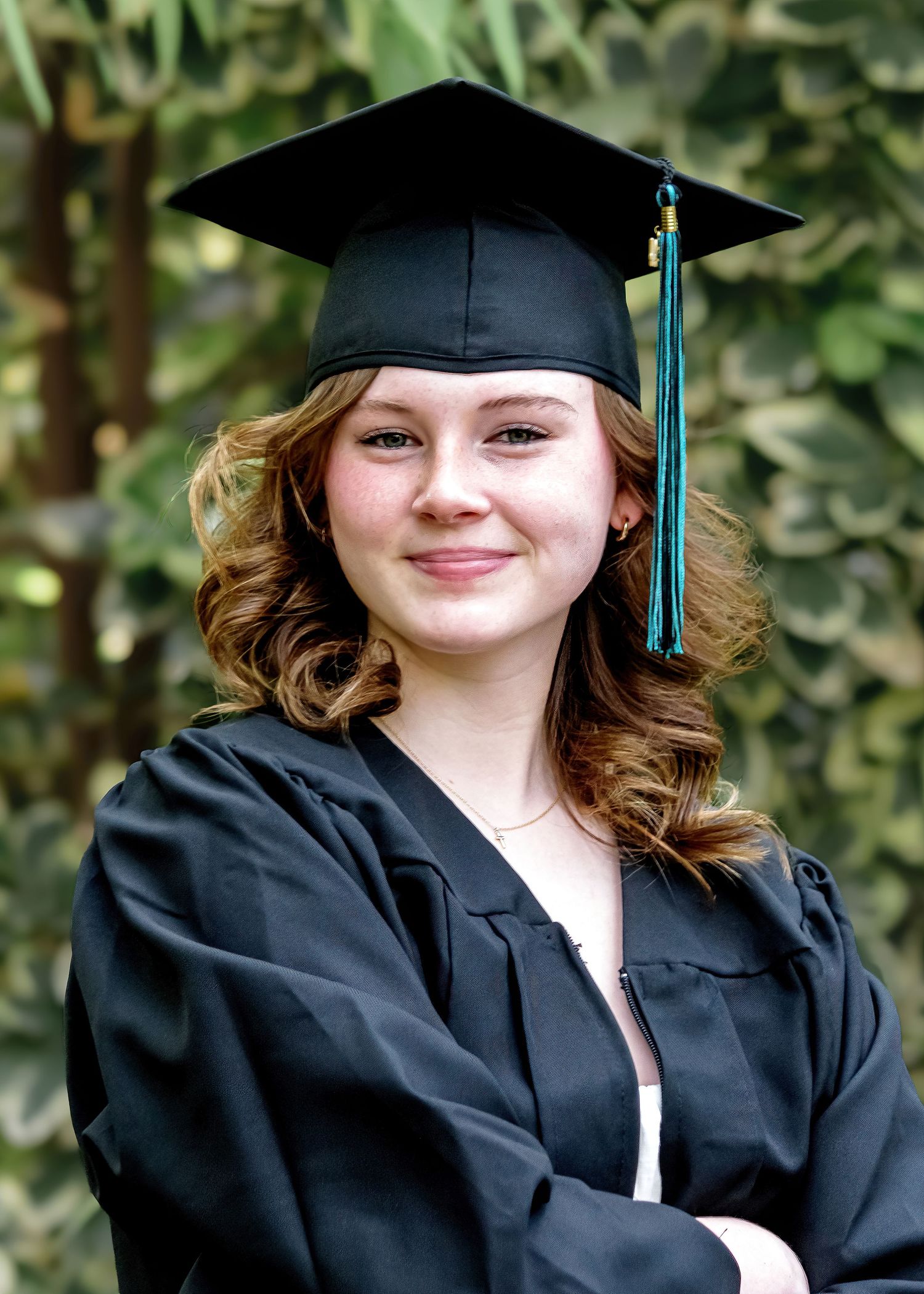 Young woman in black graduation cap and gown smiling with arms crossed in front of green foliage.