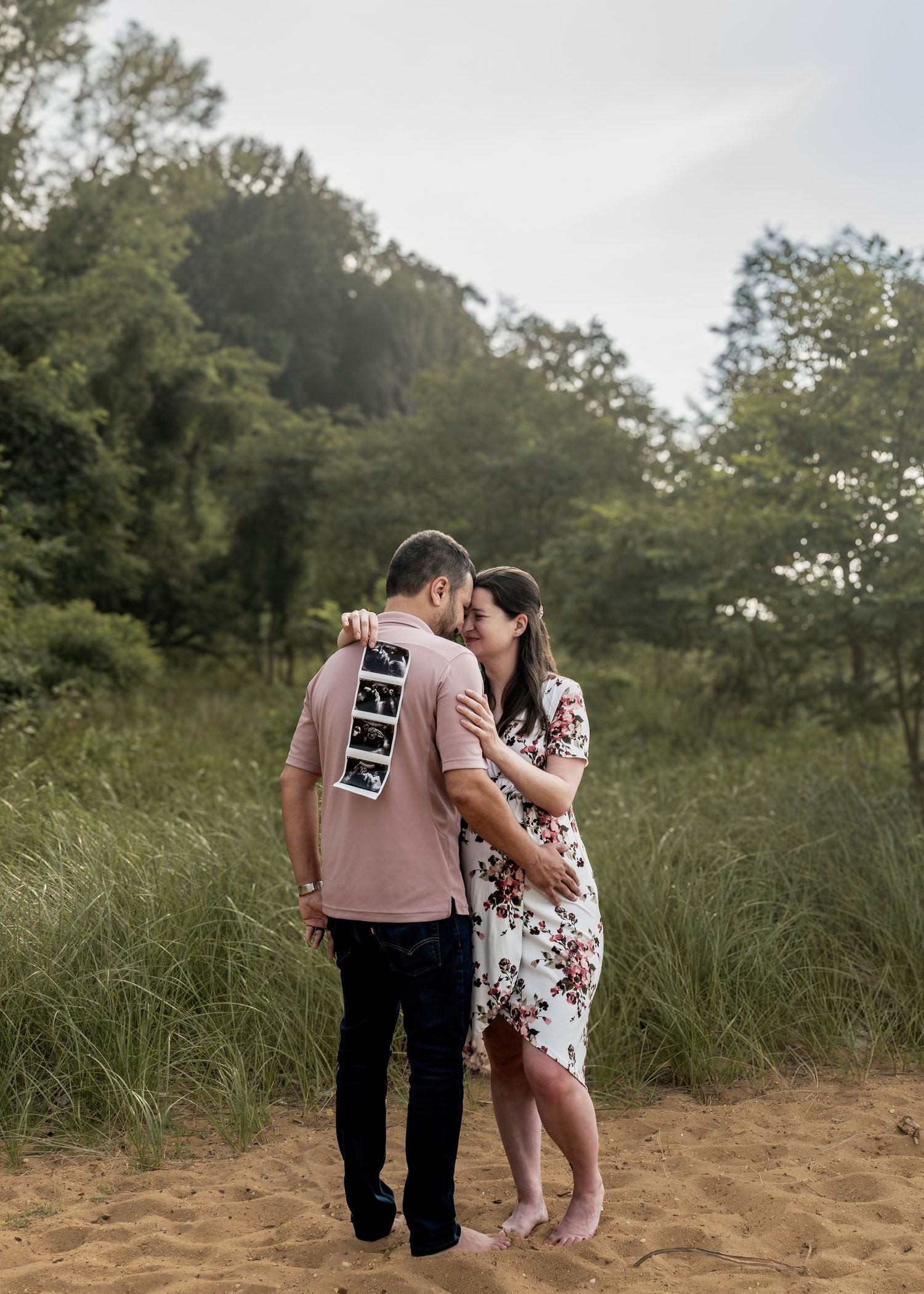 Couple sharing a kiss on sandy beach while holding ultrasound strip photos during maternity session.