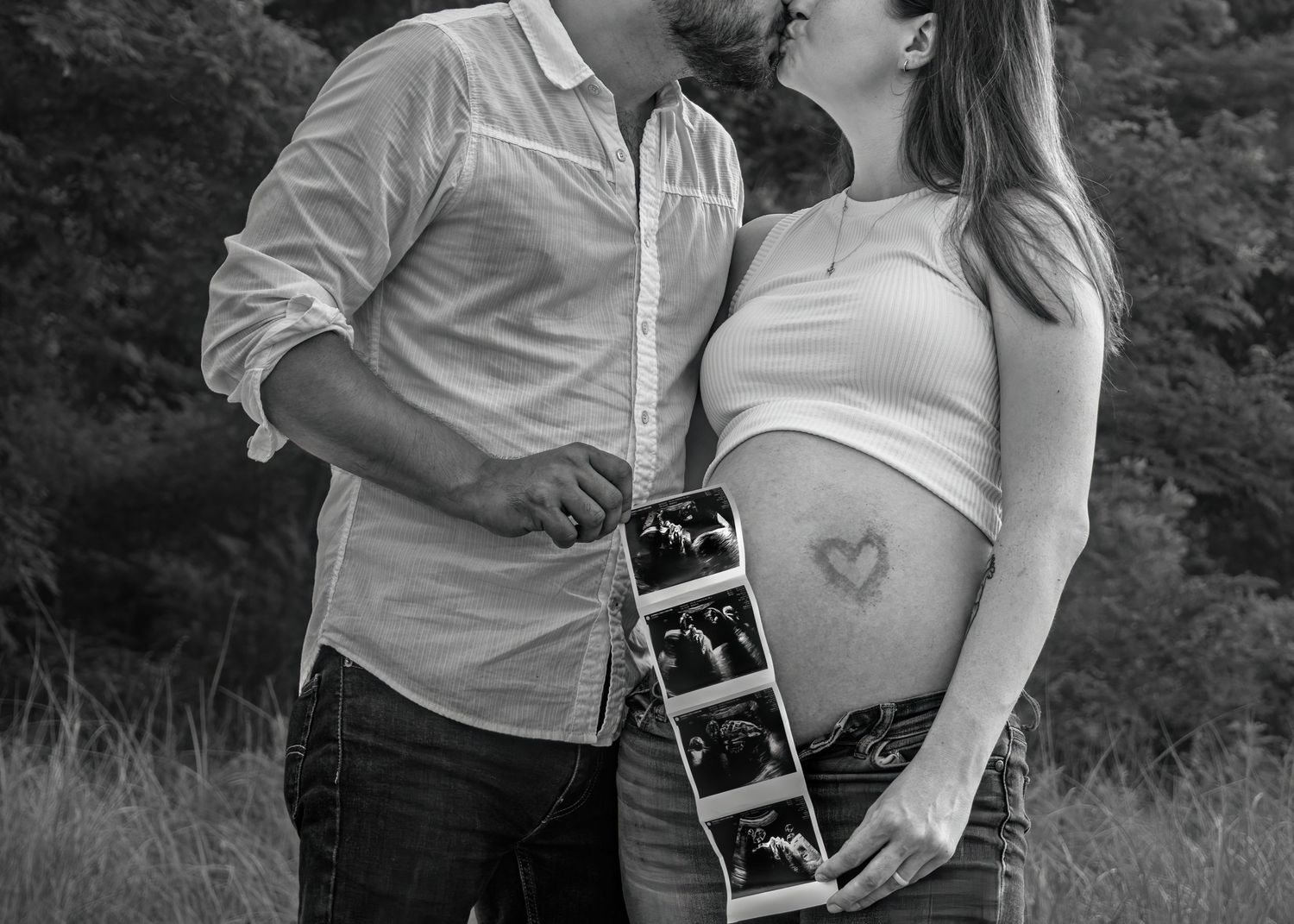 Black and white maternity photo of couple kissing holding ultrasound pictures outdoors.