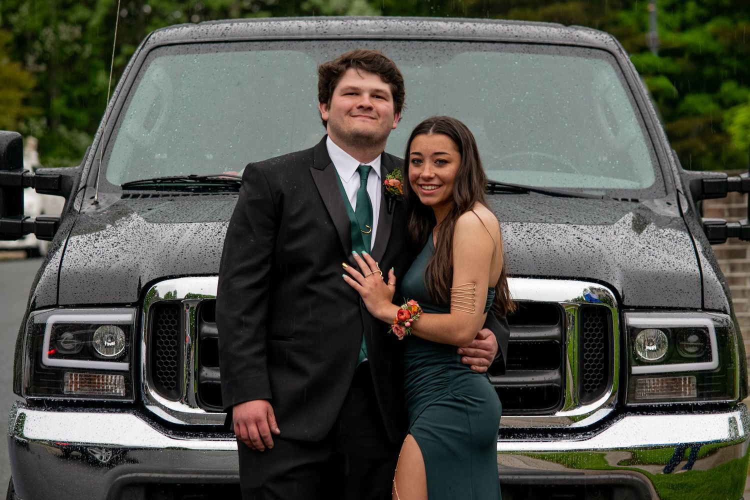 Prom couple in formal attire posing in front of a large black truck outdoors.