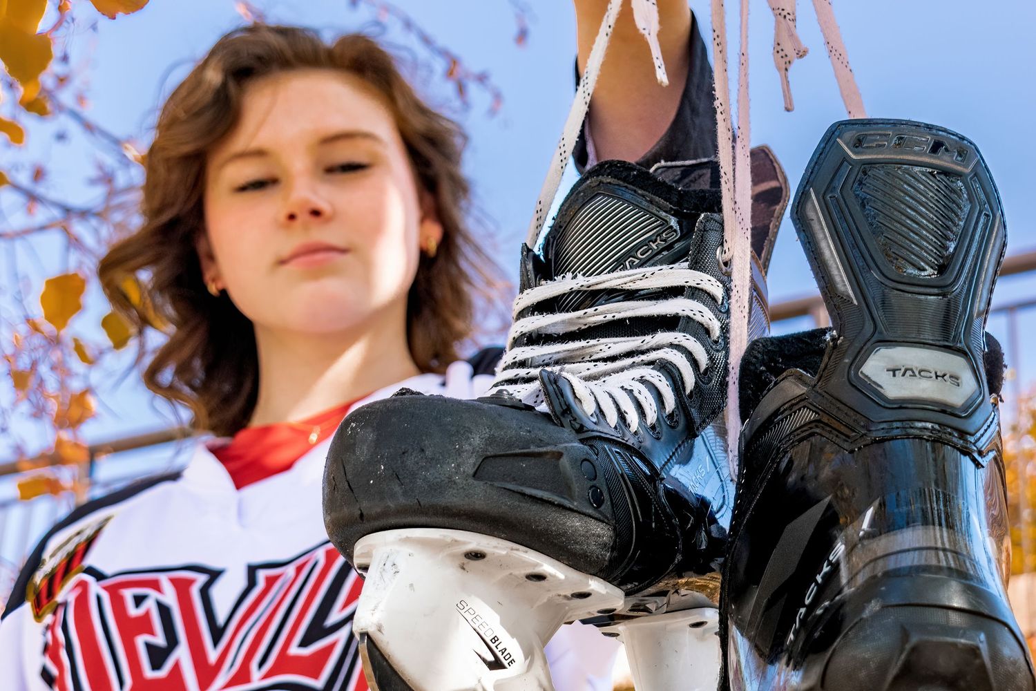 A young woman in a hockey jersey holds ice skates up close to the camera against a blue sky background.