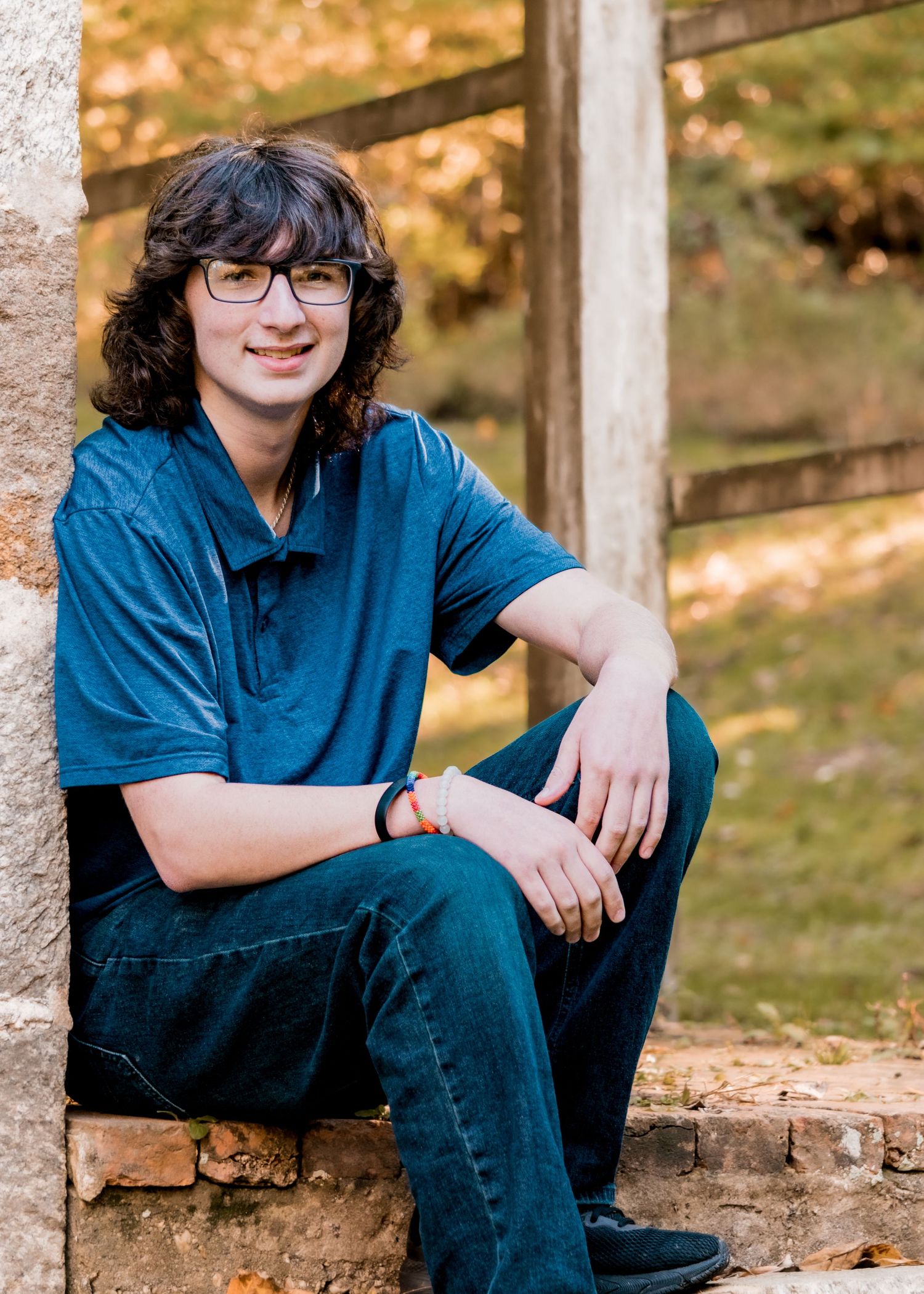 Young man with glasses and dark hair sitting on stone wall in autumn outdoor setting.