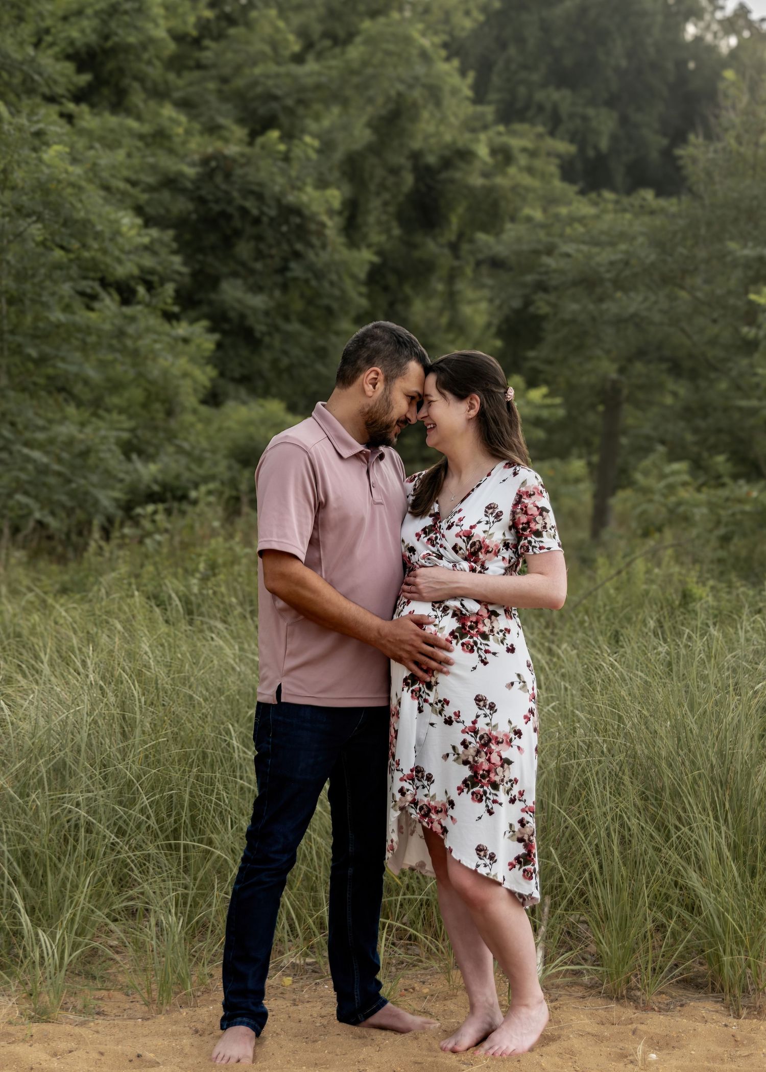 Expecting couple touching foreheads tenderly in floral dress and pink shirt outdoors.