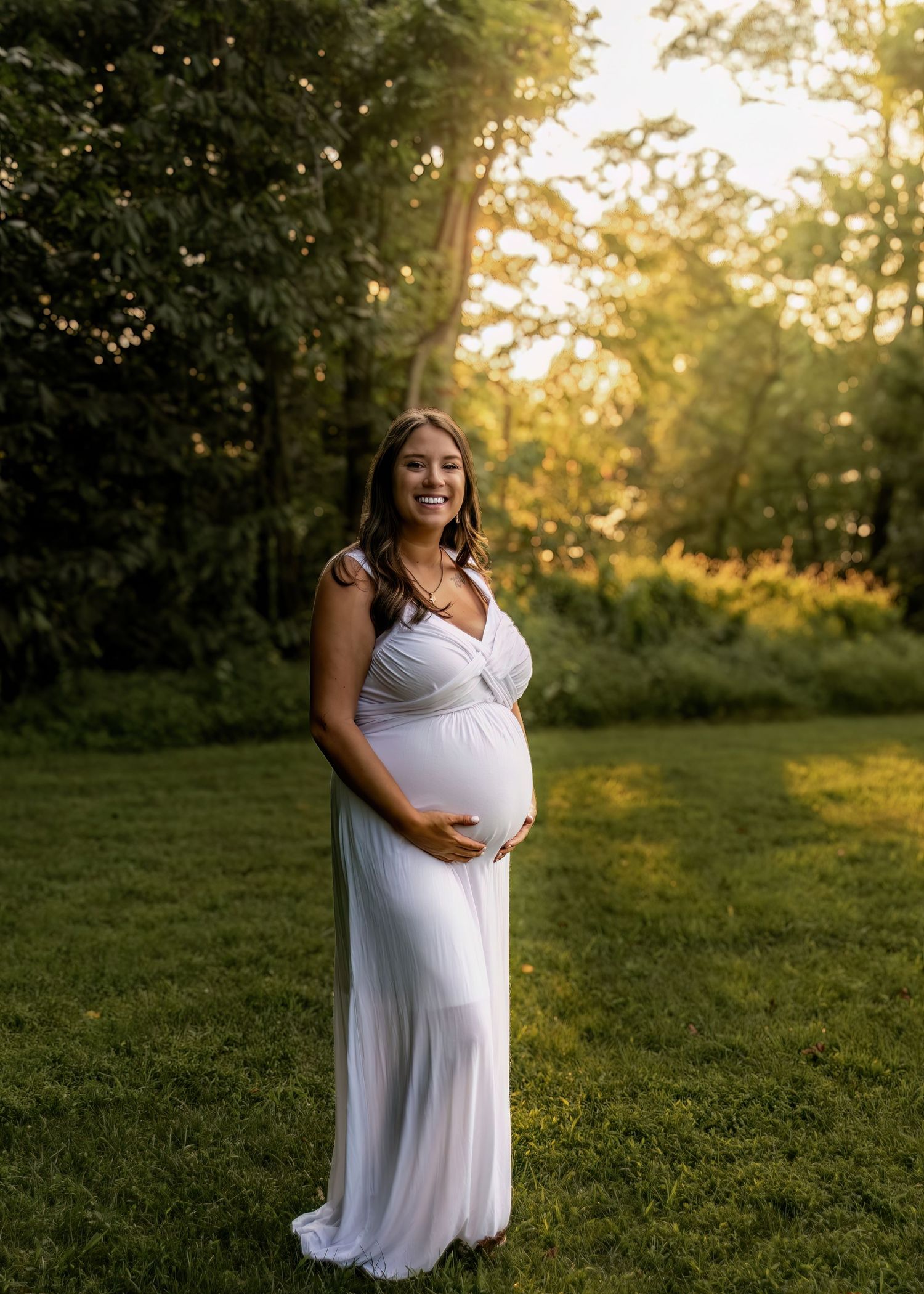 Glowing pregnant woman in white maxi dress smiling in sunlit green outdoor setting.