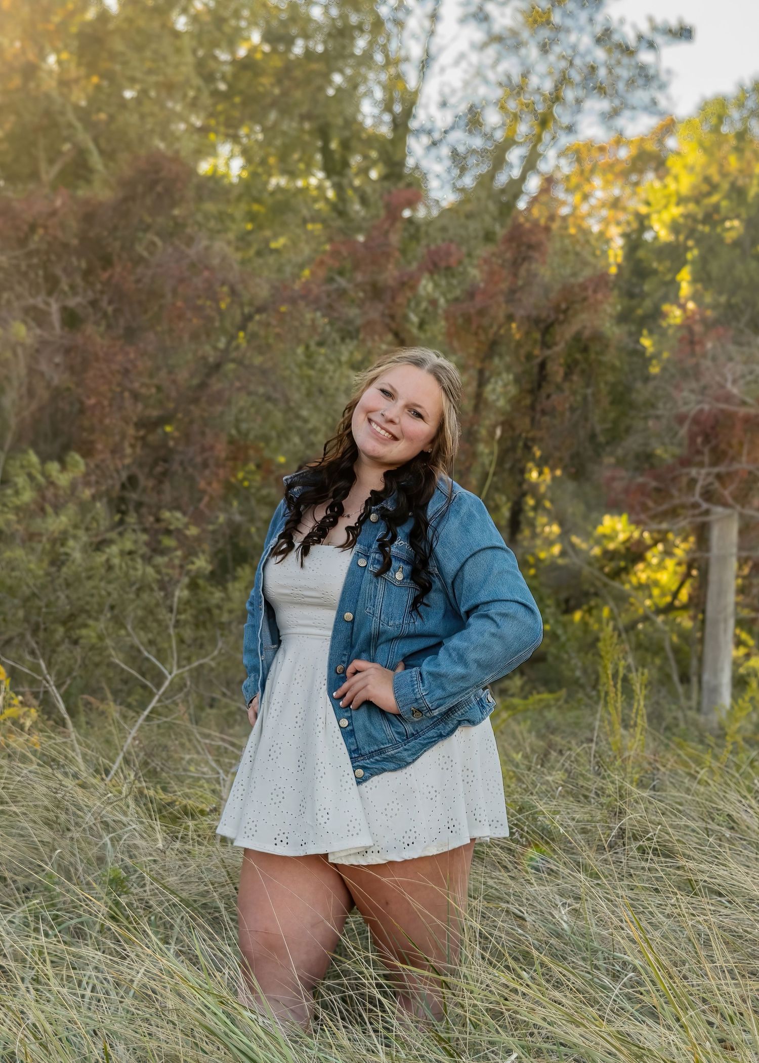 Young woman in white dress and denim jacket posing in golden autumn meadow during sunset.