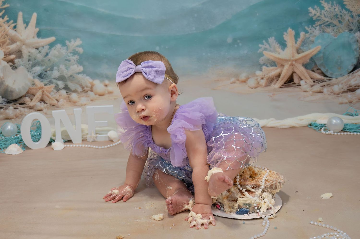Baby girl in purple tutu crawls near ONE sign at mermaid-themed first birthday cake smash photo session.