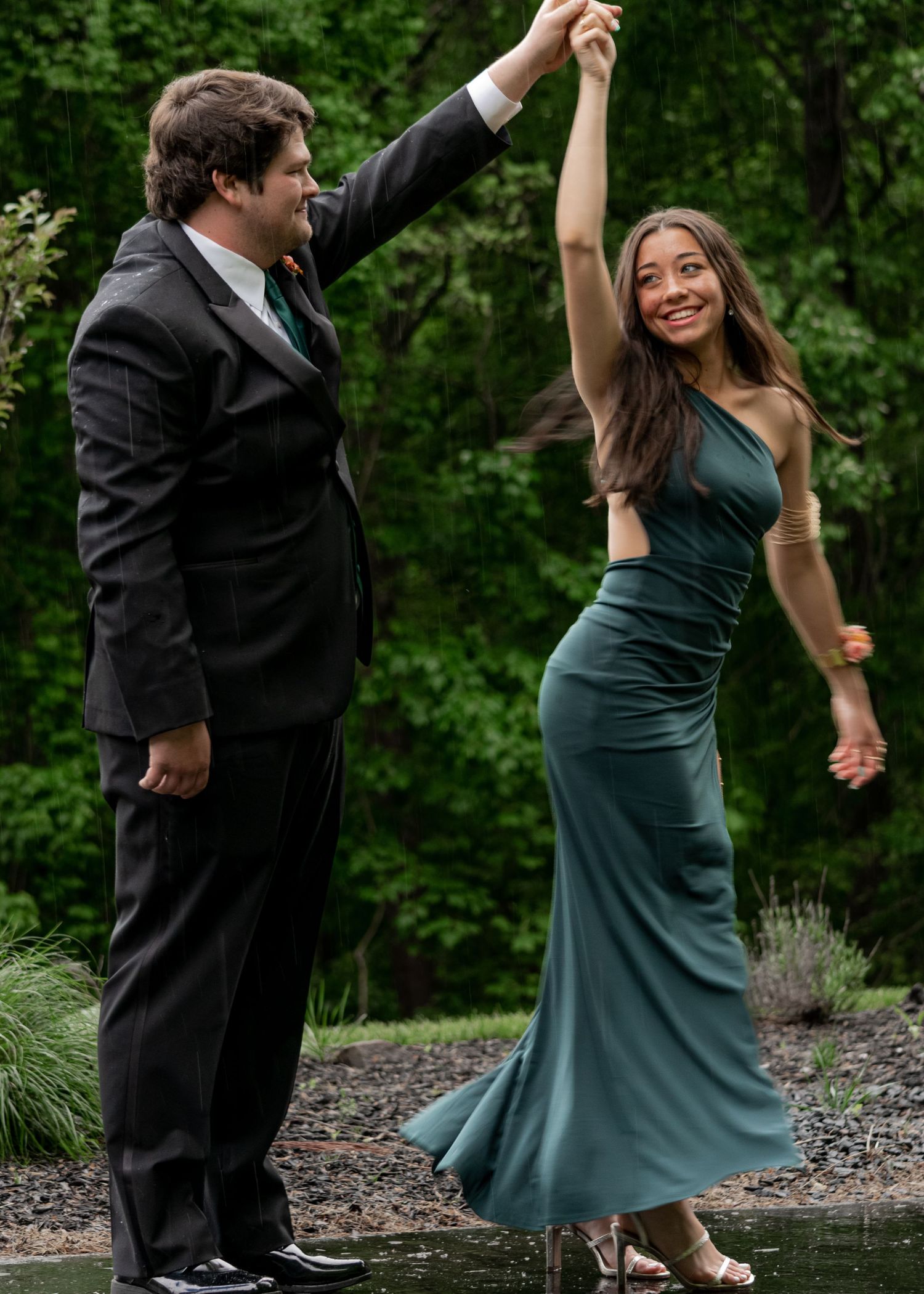 Couple dancing outdoors at prom, man in black suit spinning woman in teal green gown.