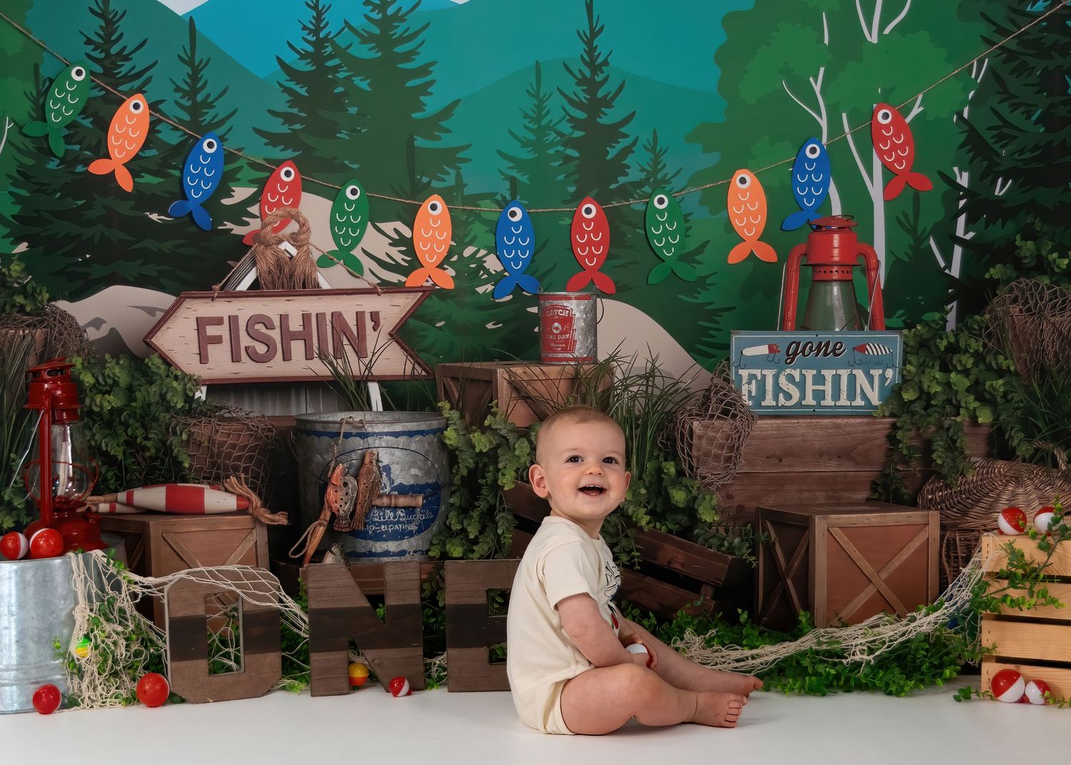 Smiling baby boy in white onesie sits at colorful fishing-themed first birthday cake smash photo session.