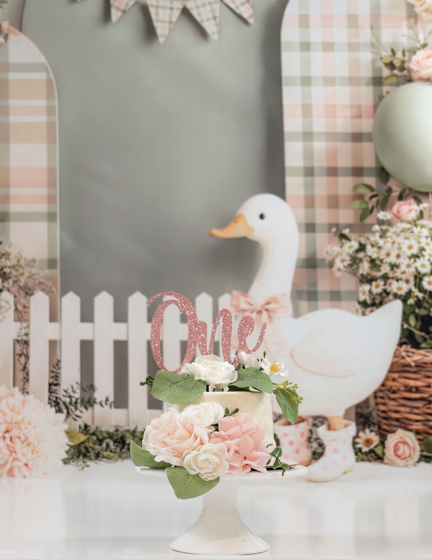 Elegant first birthday cake with pink roses and glitter One topper beside decorative duck at garden party setup.