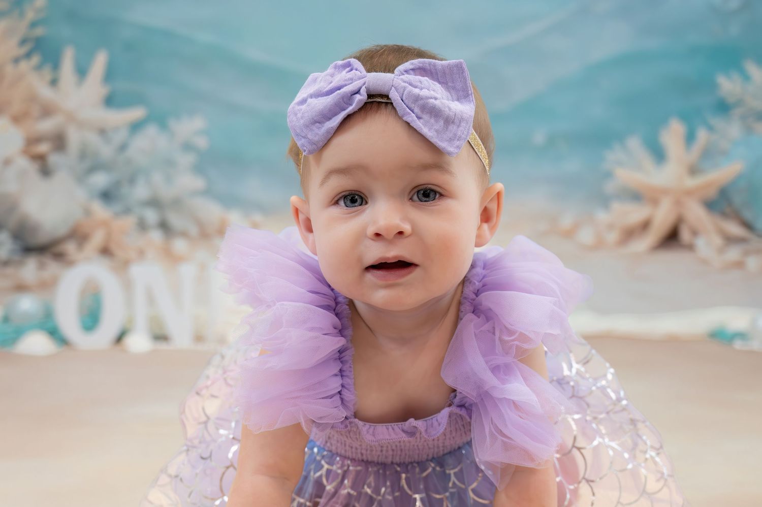 Adorable baby girl in purple tulle dress with bow headband at ocean-themed photoshoot.
