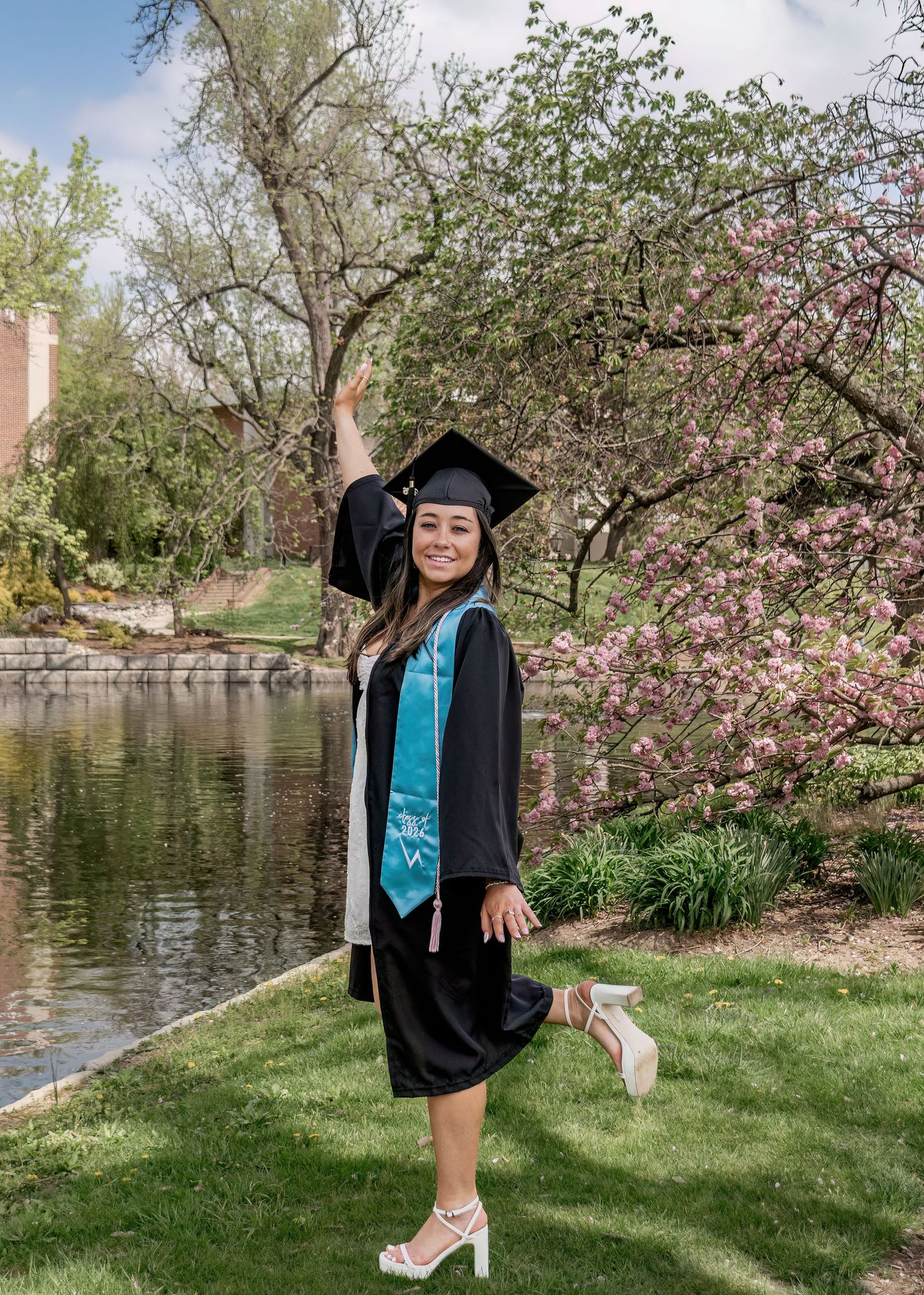 Female graduate in cap and gown posing near pond with blooming pink trees in spring.