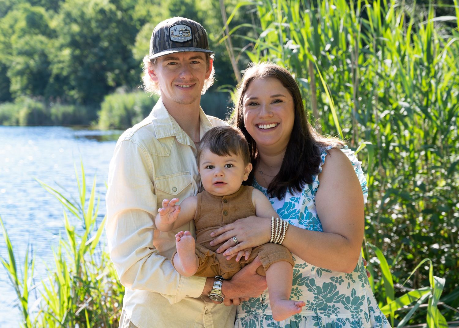 Young family with baby posing by lake surrounded by tall green reeds.