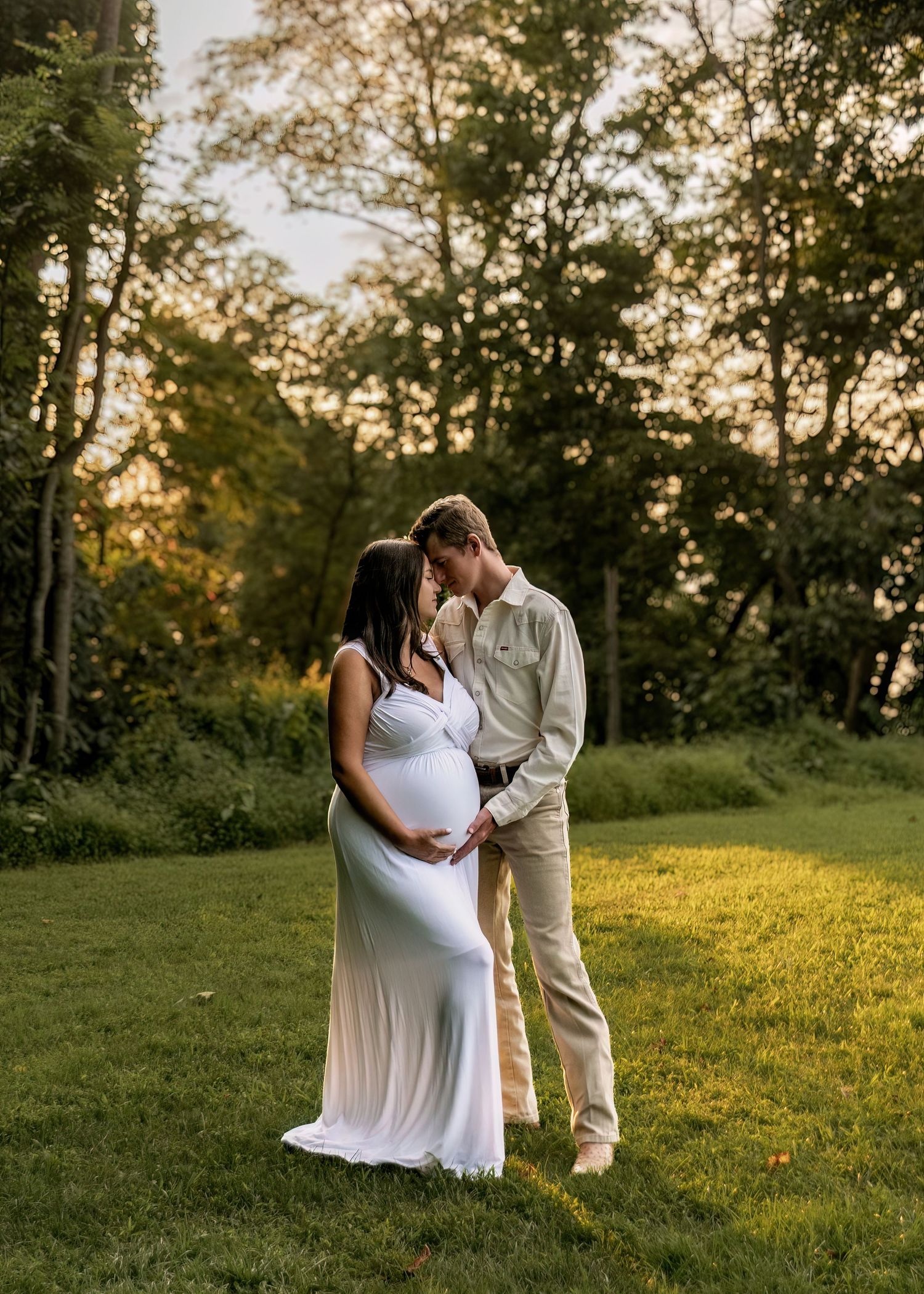 Pregnant couple sharing intimate moment at golden hour on lush green lawn outdoors.