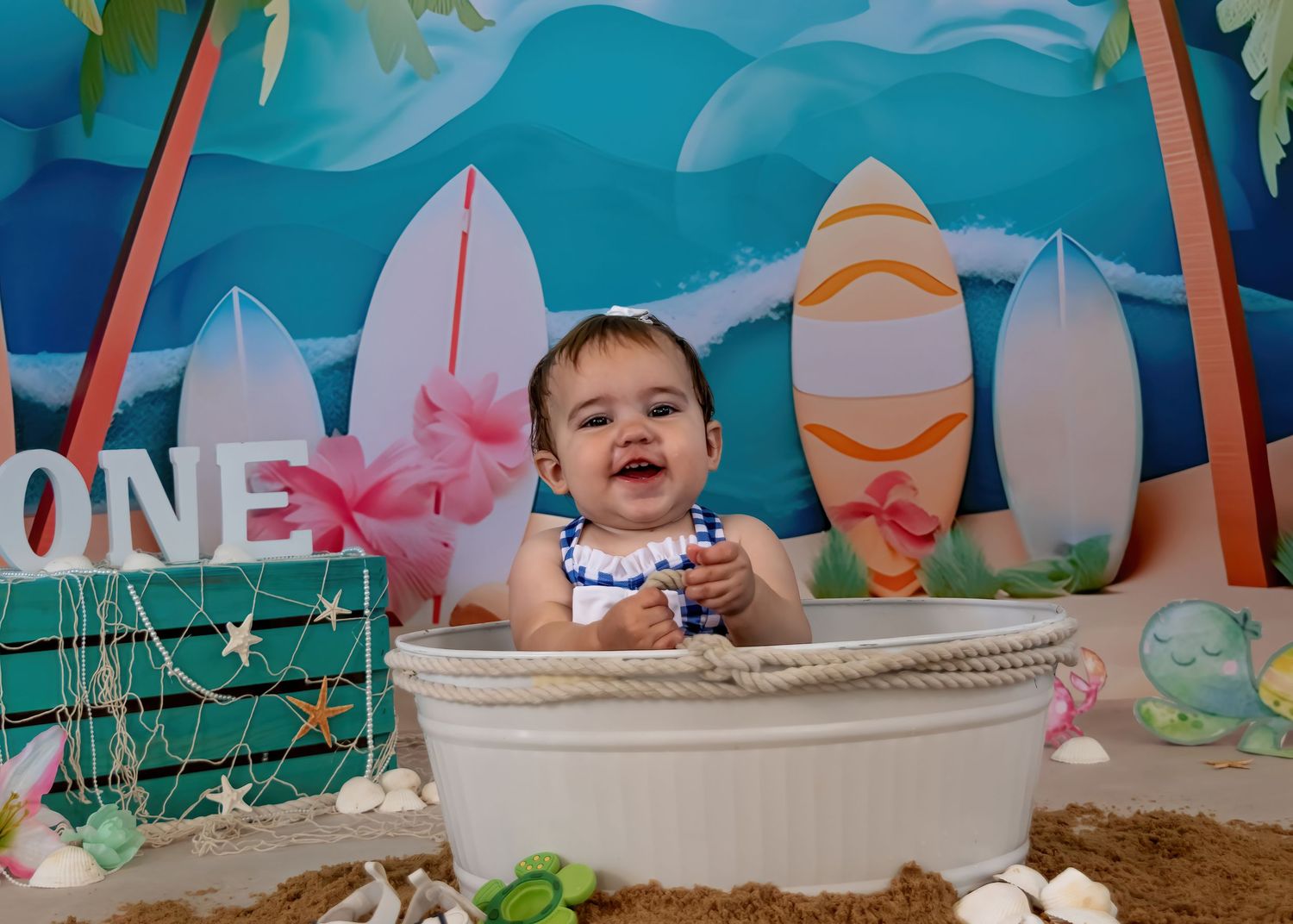 Laughing baby girl sits in white tub at beach-themed first birthday session with surfboards backdrop.