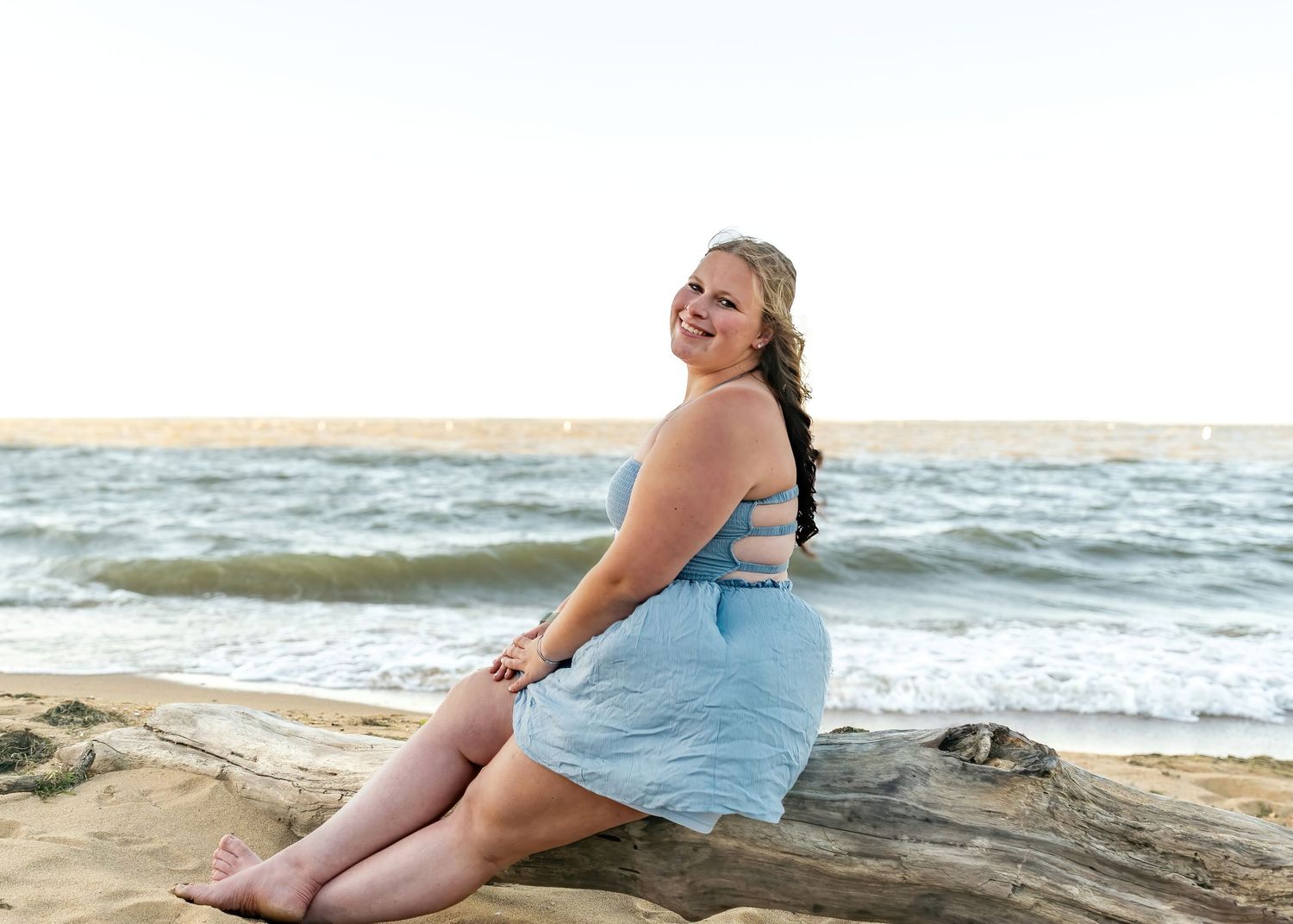 Smiling young woman in light blue dress sitting on driftwood at a sunny beach.