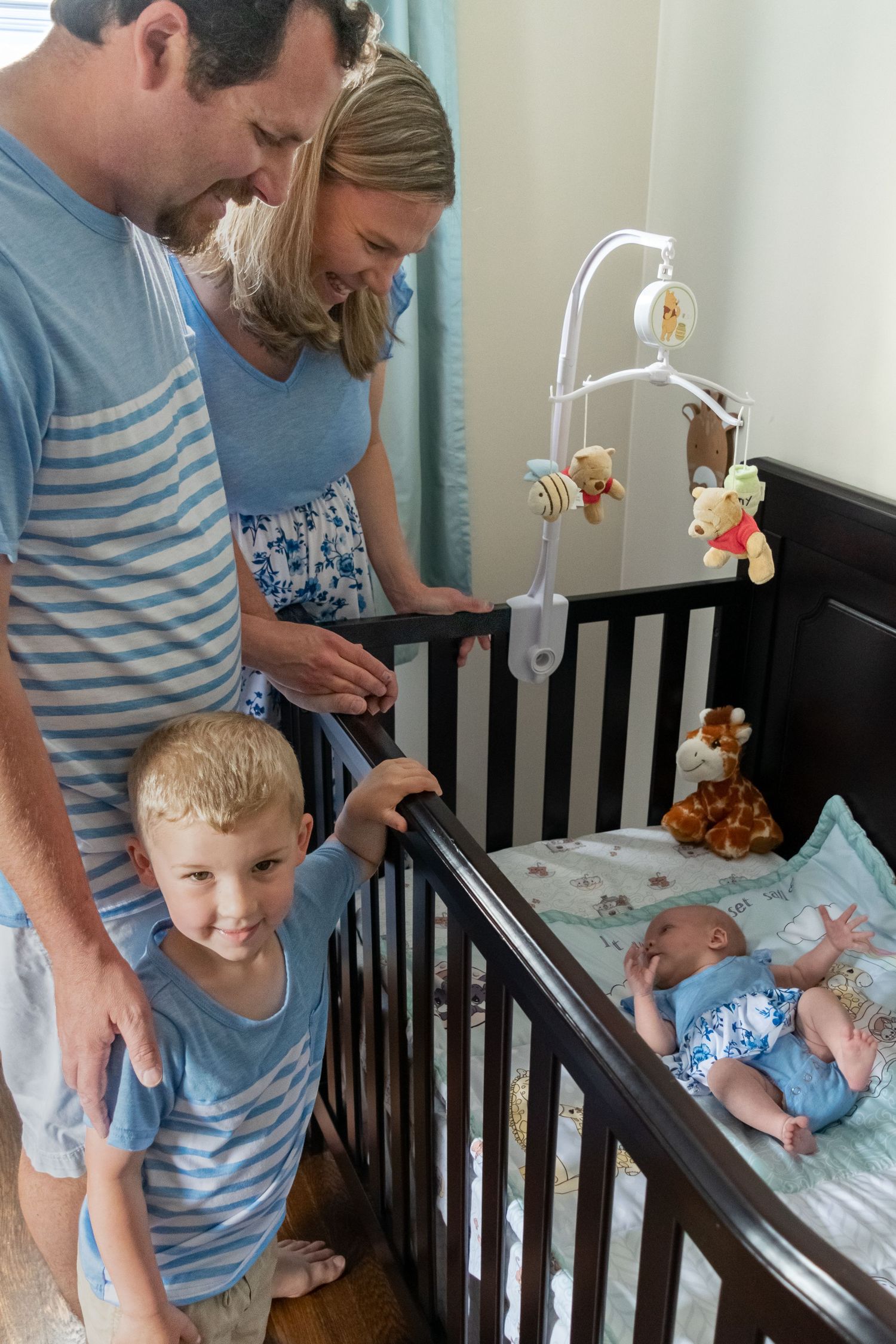 Happy family of four with toddler son smiling at newborn baby in dark wood crib.