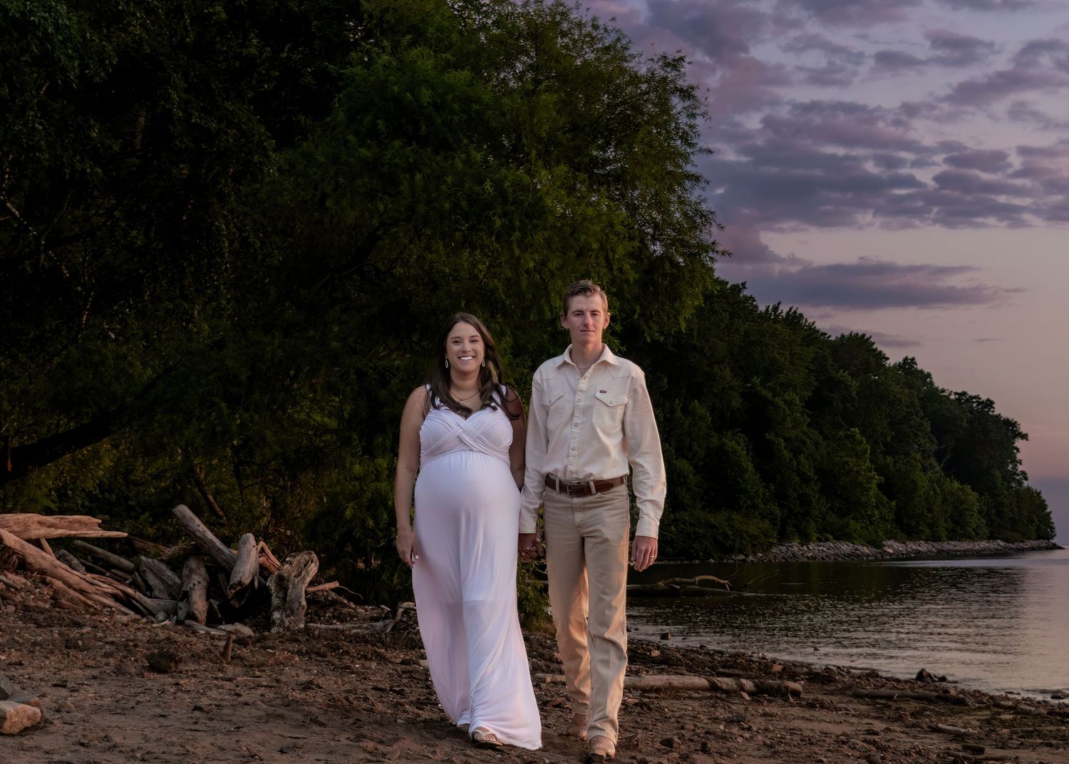 Pregnant couple walking hand in hand along a scenic beach at sunset during maternity photoshoot.