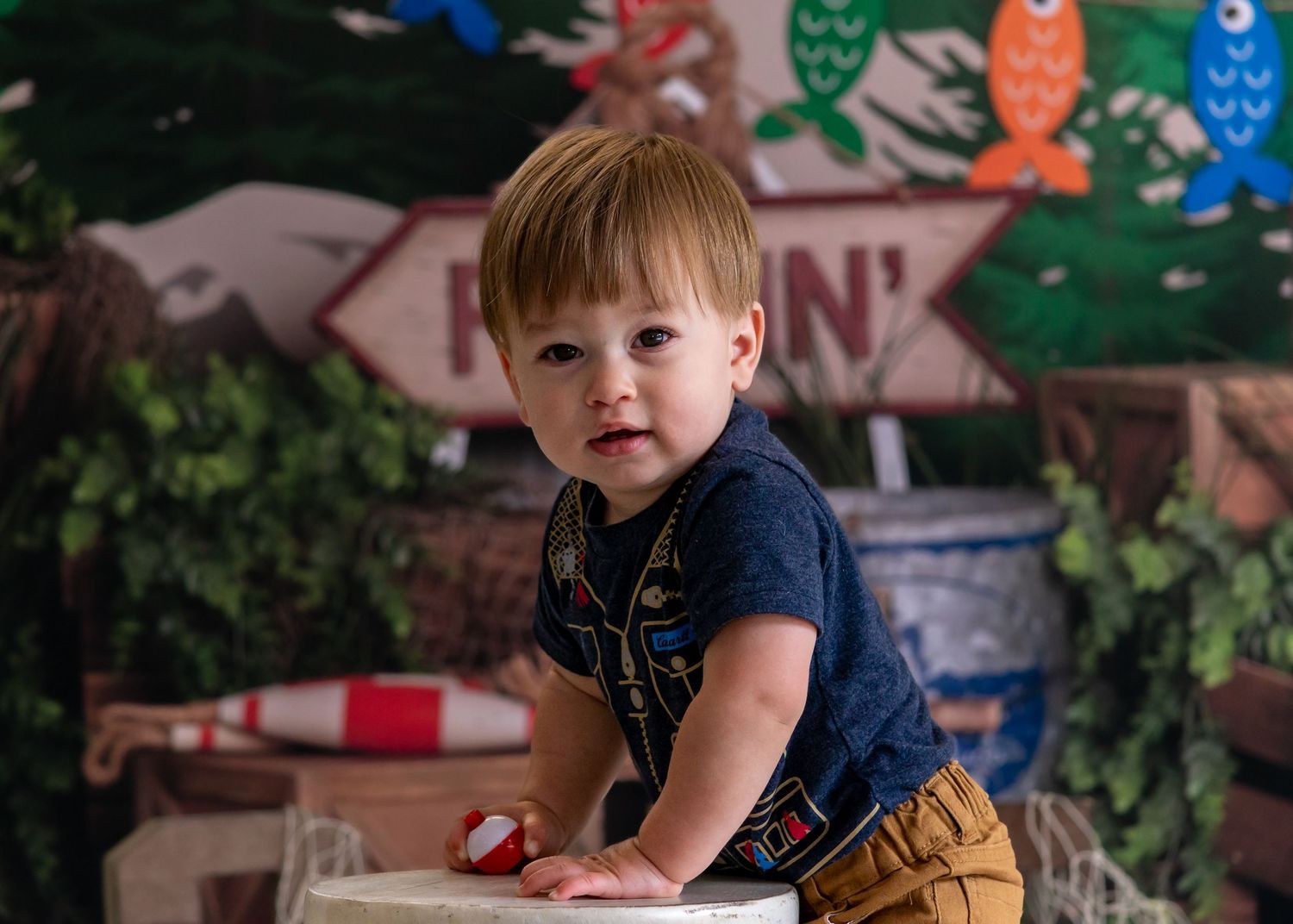Adorable toddler boy in navy outfit leans on white stool at fishing-themed birthday photo session.