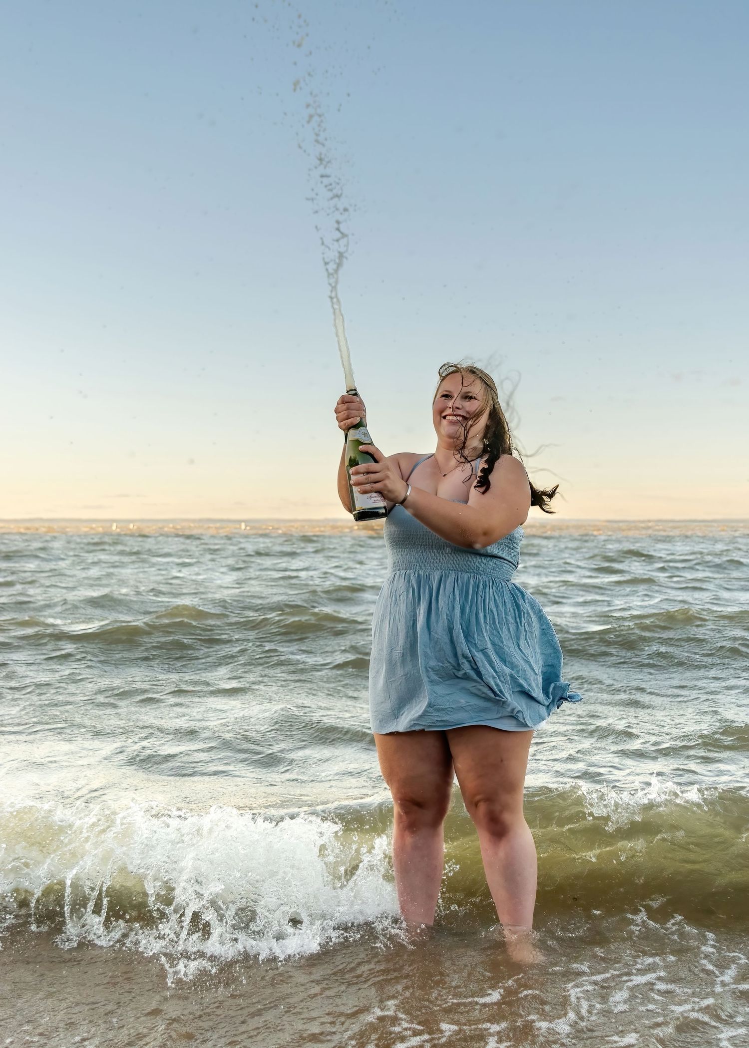 Young woman celebrating on beach at sunset popping champagne bottle with ocean waves behind her.