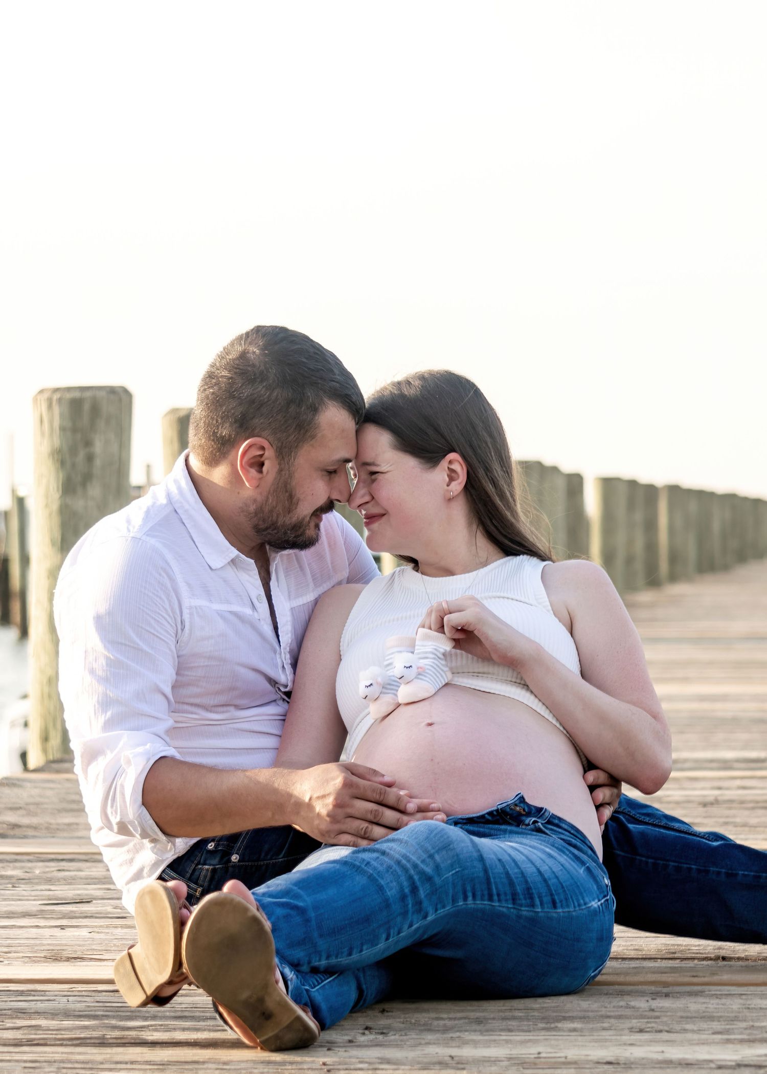 Expecting couple sitting on wooden dock, smiling at each other while holding tiny baby shoes on bump.