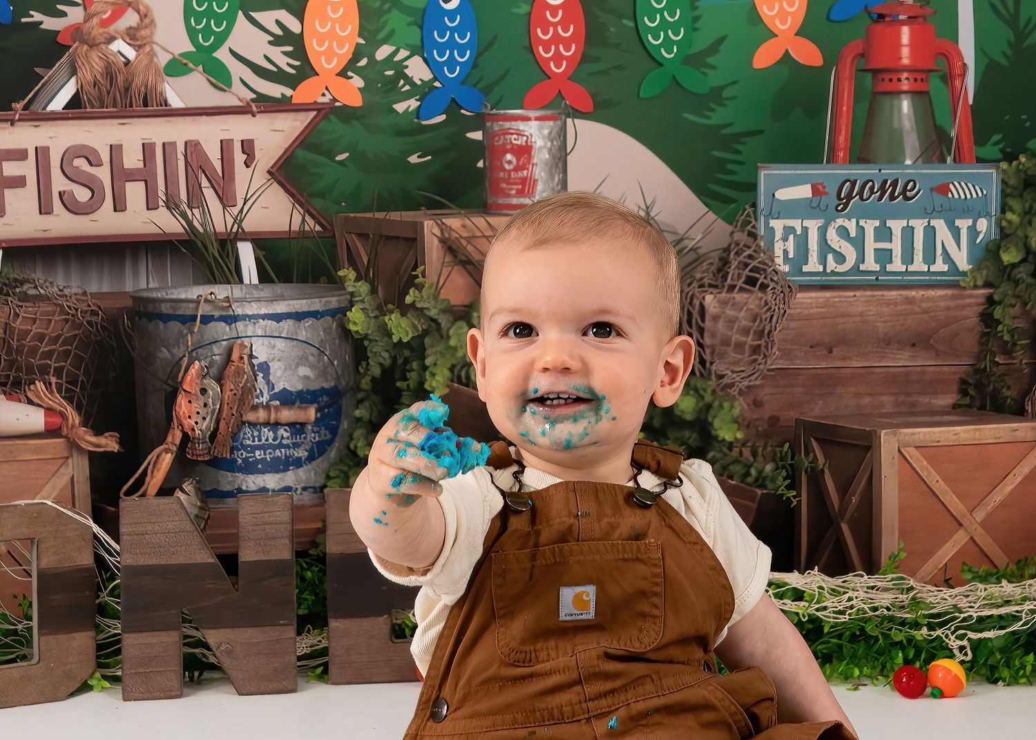 Baby boy in overalls celebrates first birthday with fishing theme cake smash, smiling with blue frosting on face.