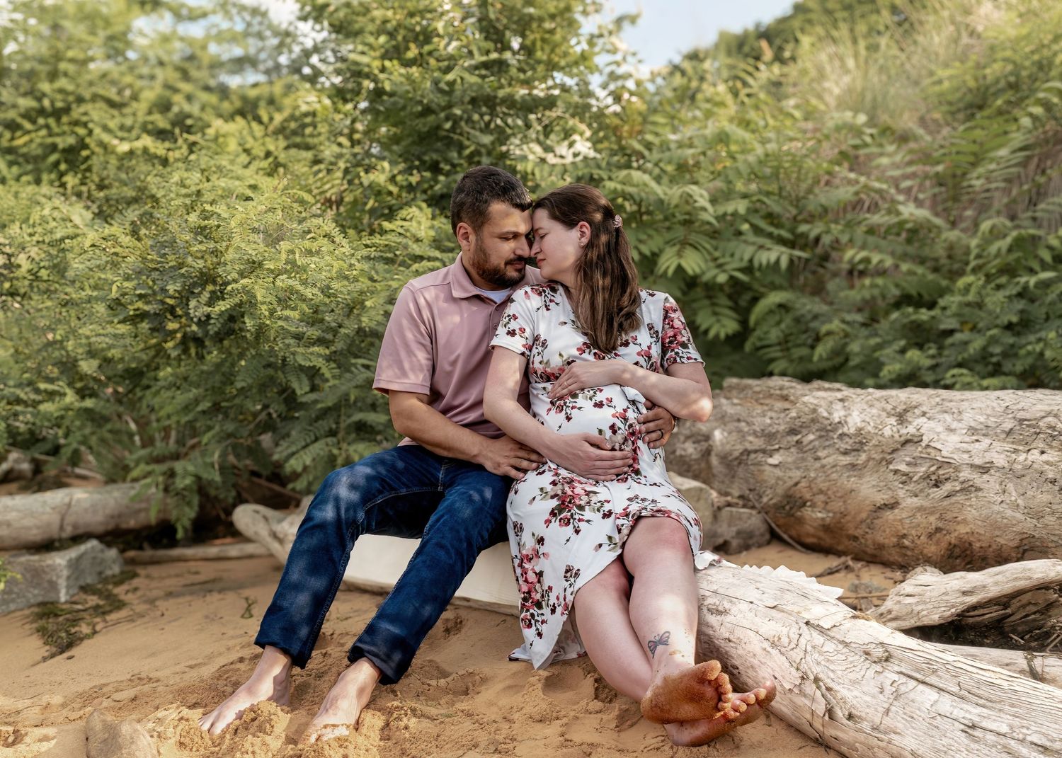 Happy expecting couple sitting on driftwood logs, smiling at each other during outdoor maternity session.