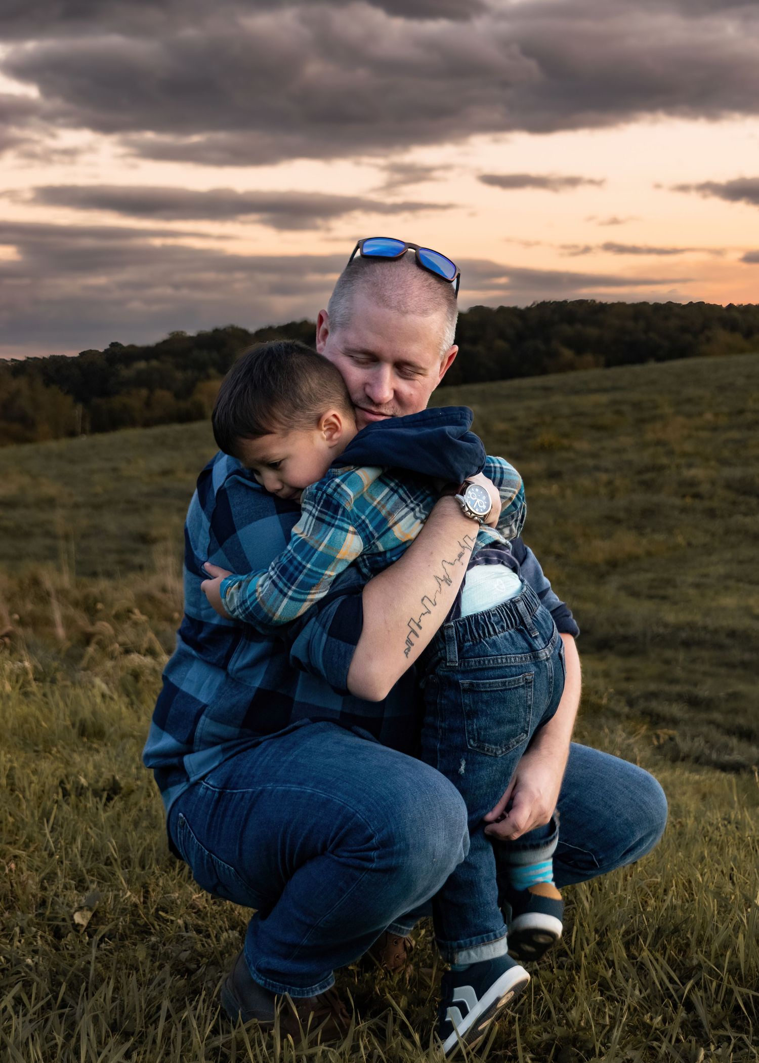 Bald man hugging young boy in field at sunset with dramatic cloudy sky.