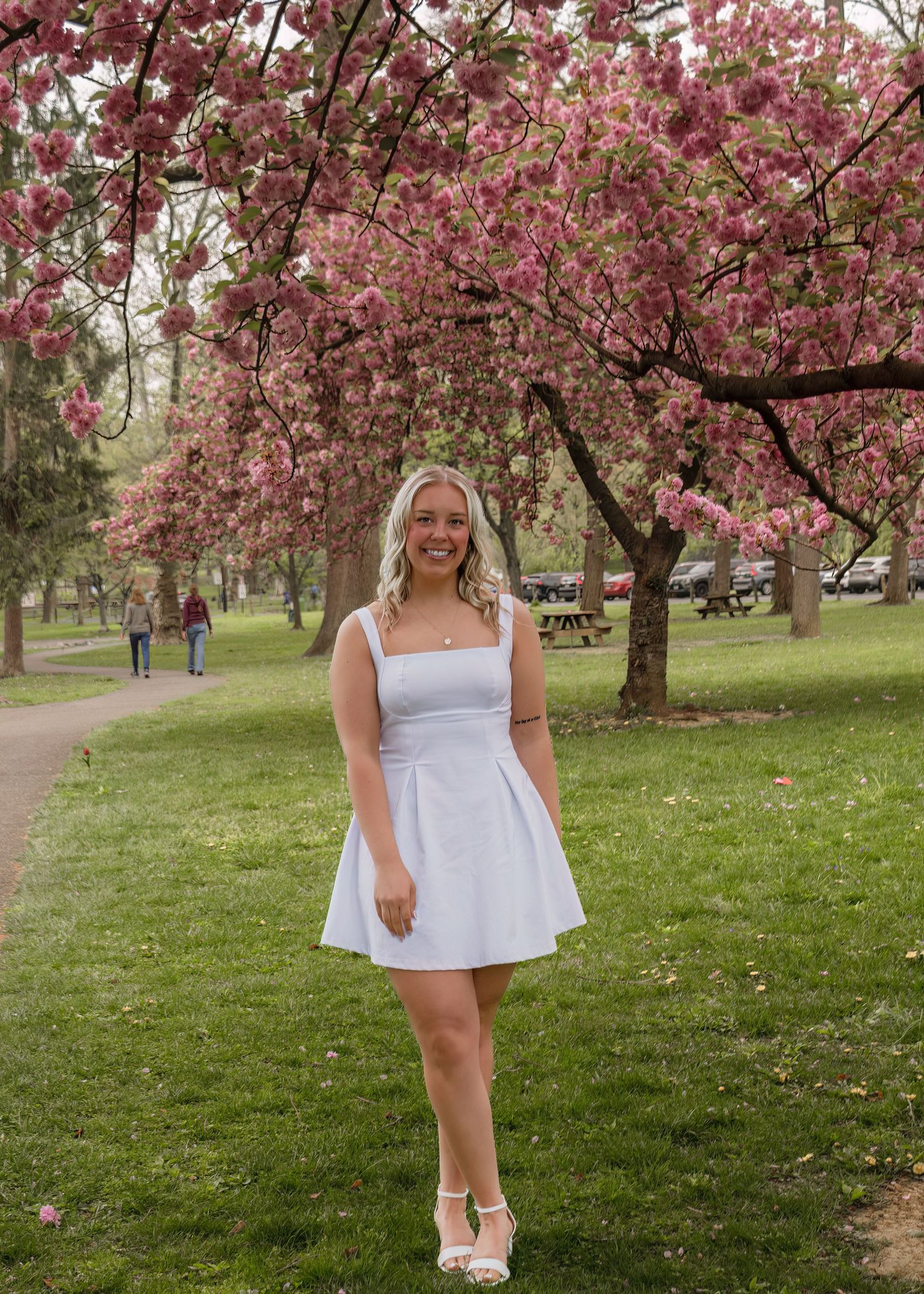A smiling blonde woman in a white mini dress poses under blooming pink cherry blossom trees in a green park.